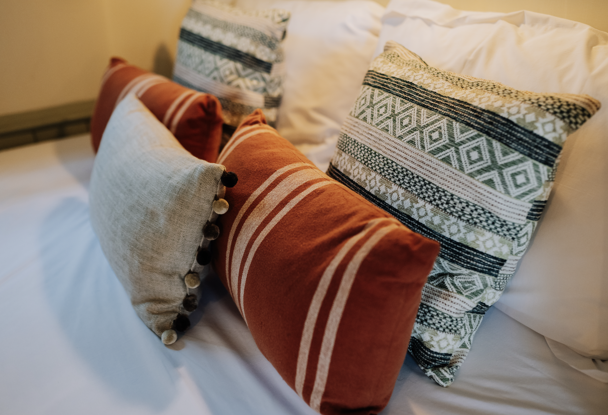 Decorative pillows on a bed with white sheets and a beige wall in the background.