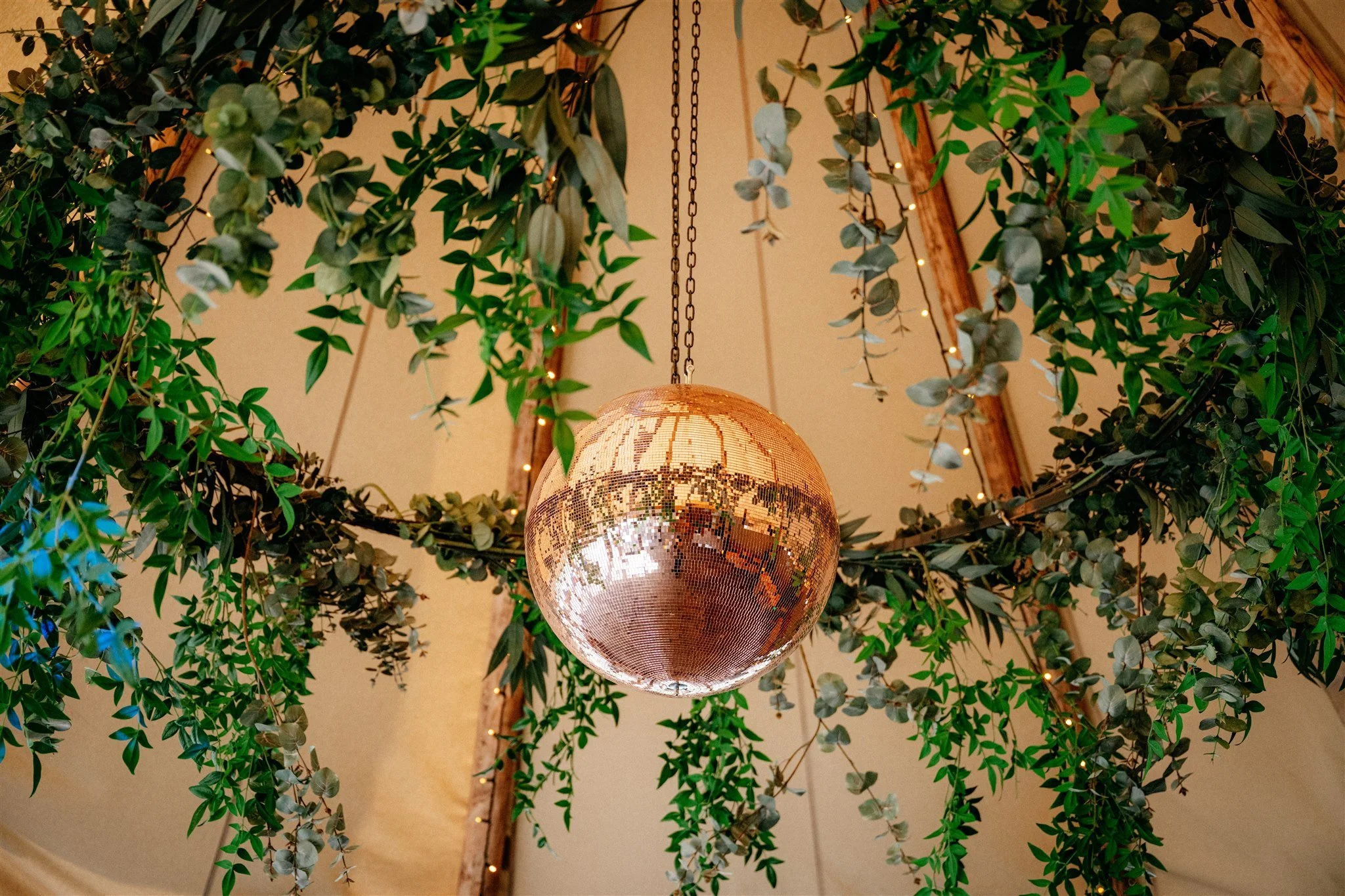 A spherical disco ball hanging from a chain surrounded by green leafy plants and string lights.