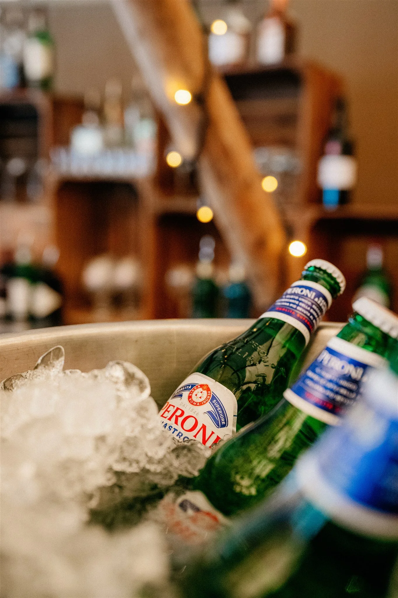 Close-up of a bowl of ice with green glass bottles, beer labels, and syrup bottles on top in the background, slightly out of focus.