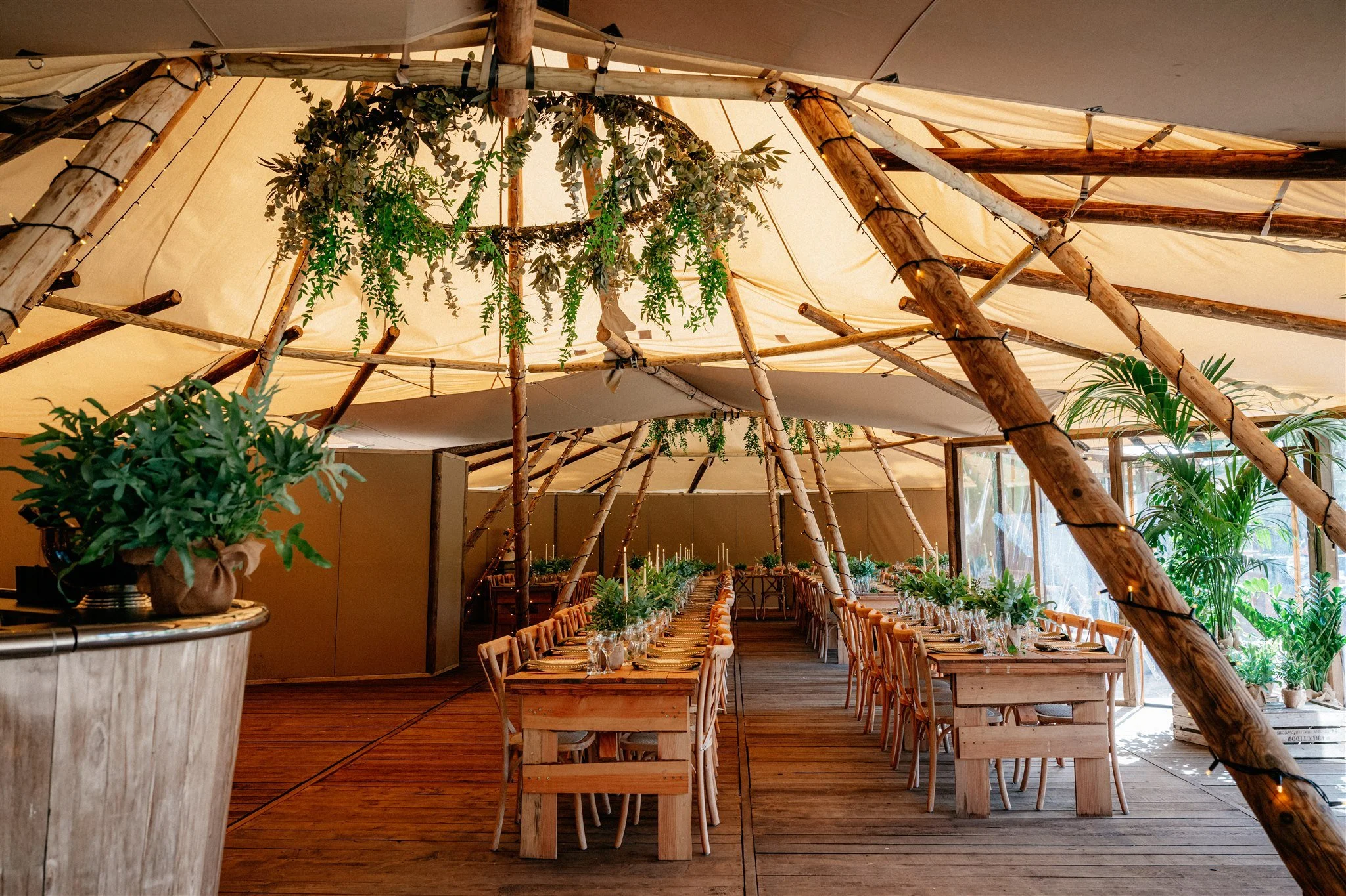 Indoor event space decorated with wooden beams, hanging greenery, potted plants, and a long dining table set with chairs and wine glasses.