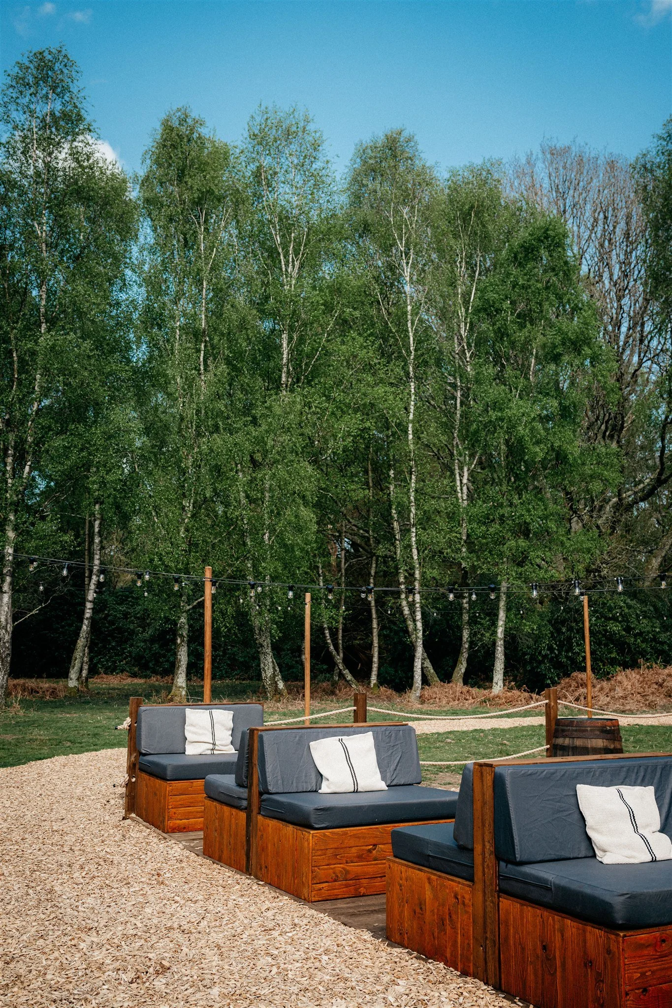 Outdoor seating area with wooden benches and dark cushions, set on gravel, with string lights and a backdrop of green trees and a blue sky.
