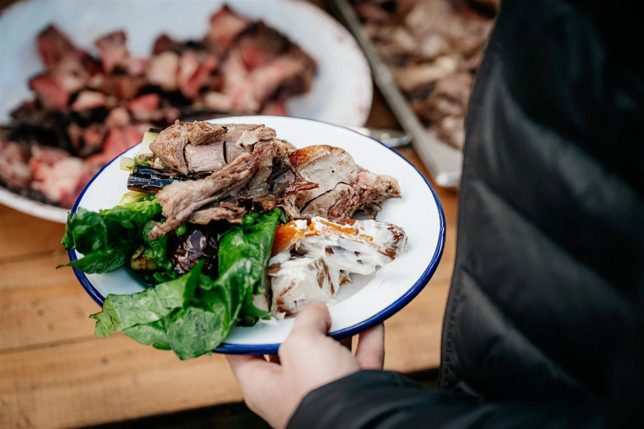 A person holding a white plate filled with cooked meats and a green salad, with grilled meat and a chopped meat dish in the background.