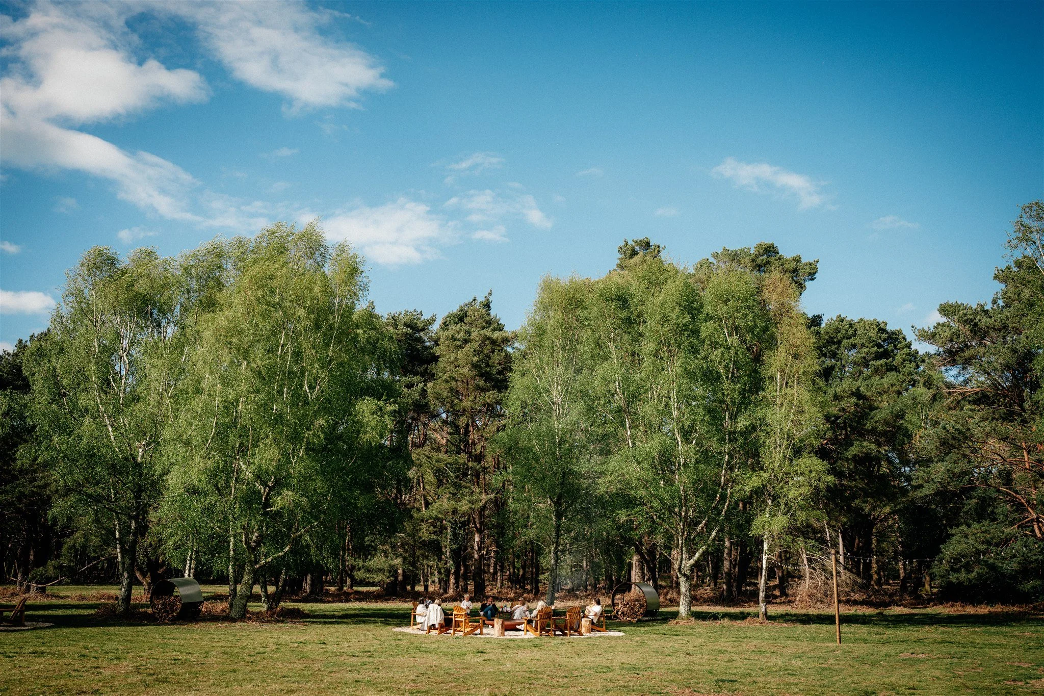 A group of people sitting on chairs around a fire pit in a park, with tall trees and blue sky with clouds in the background.