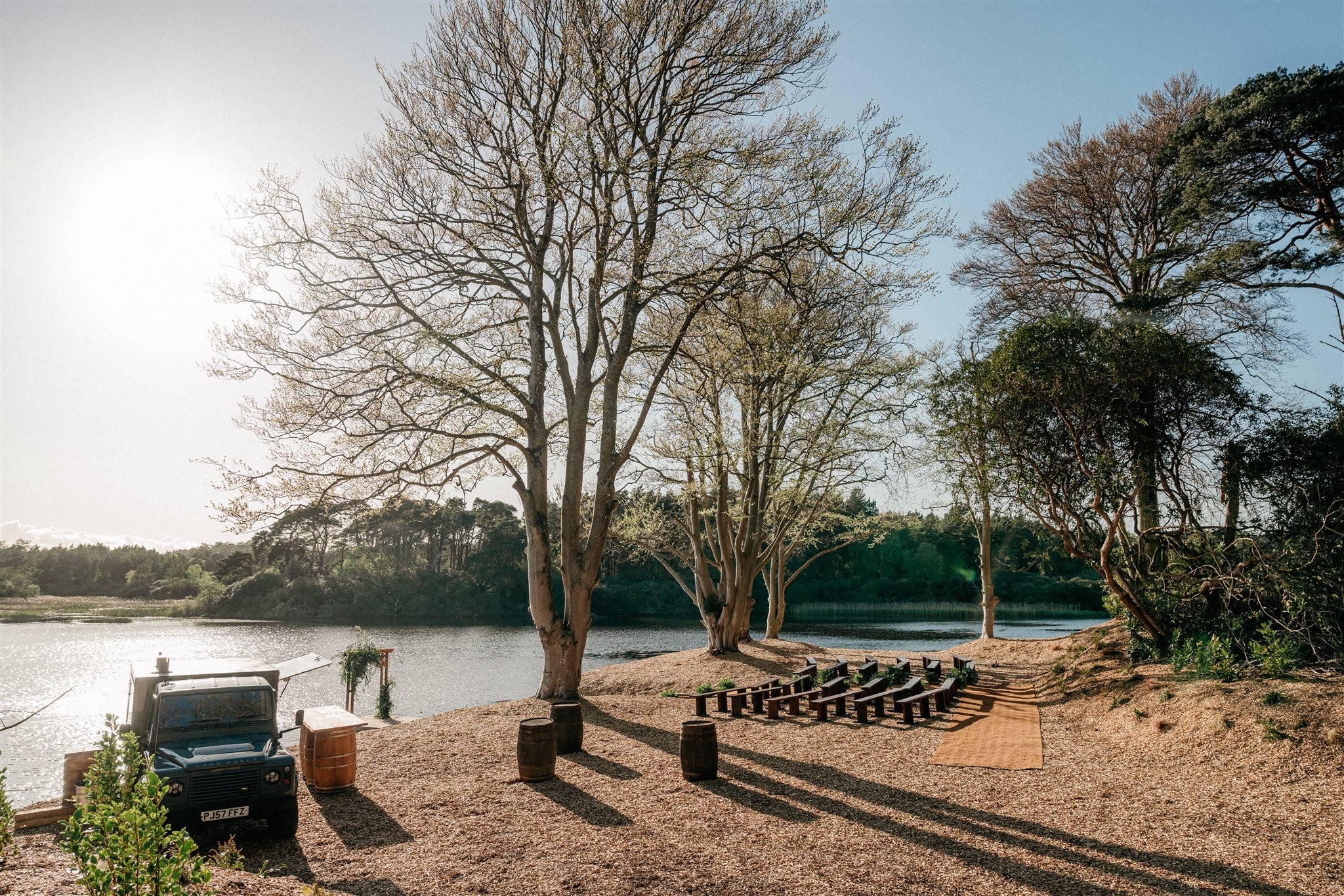 A lakeside park with several tall, leafless trees, a gravel path with benches, a boat near the water, and the sun shining in a clear blue sky.