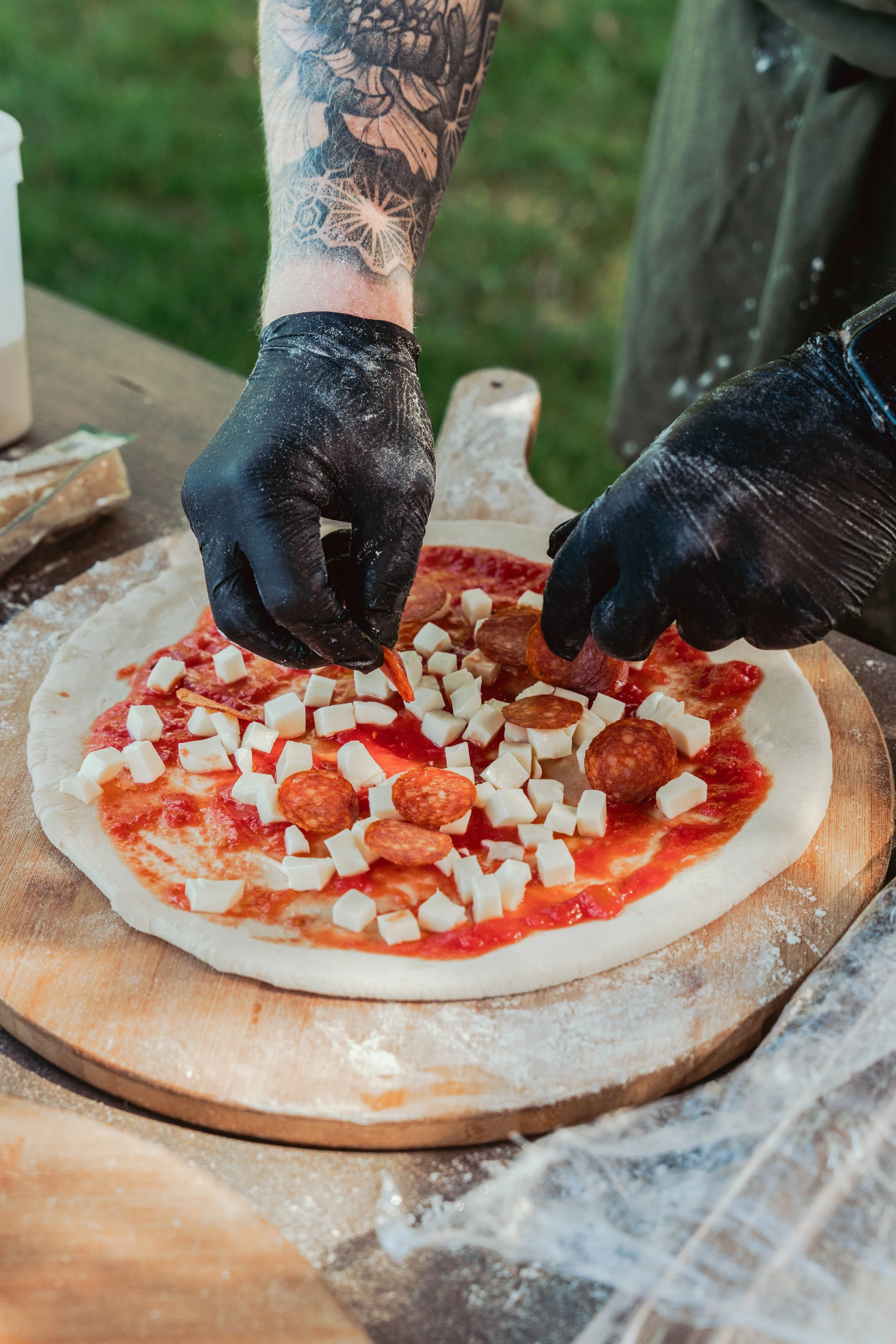 Person with tattoos on arm making a pizza, adding slices of pepperoni, cubes of cheese, and pepperoni slices to the dough on a wooden pizza peel outdoors.
