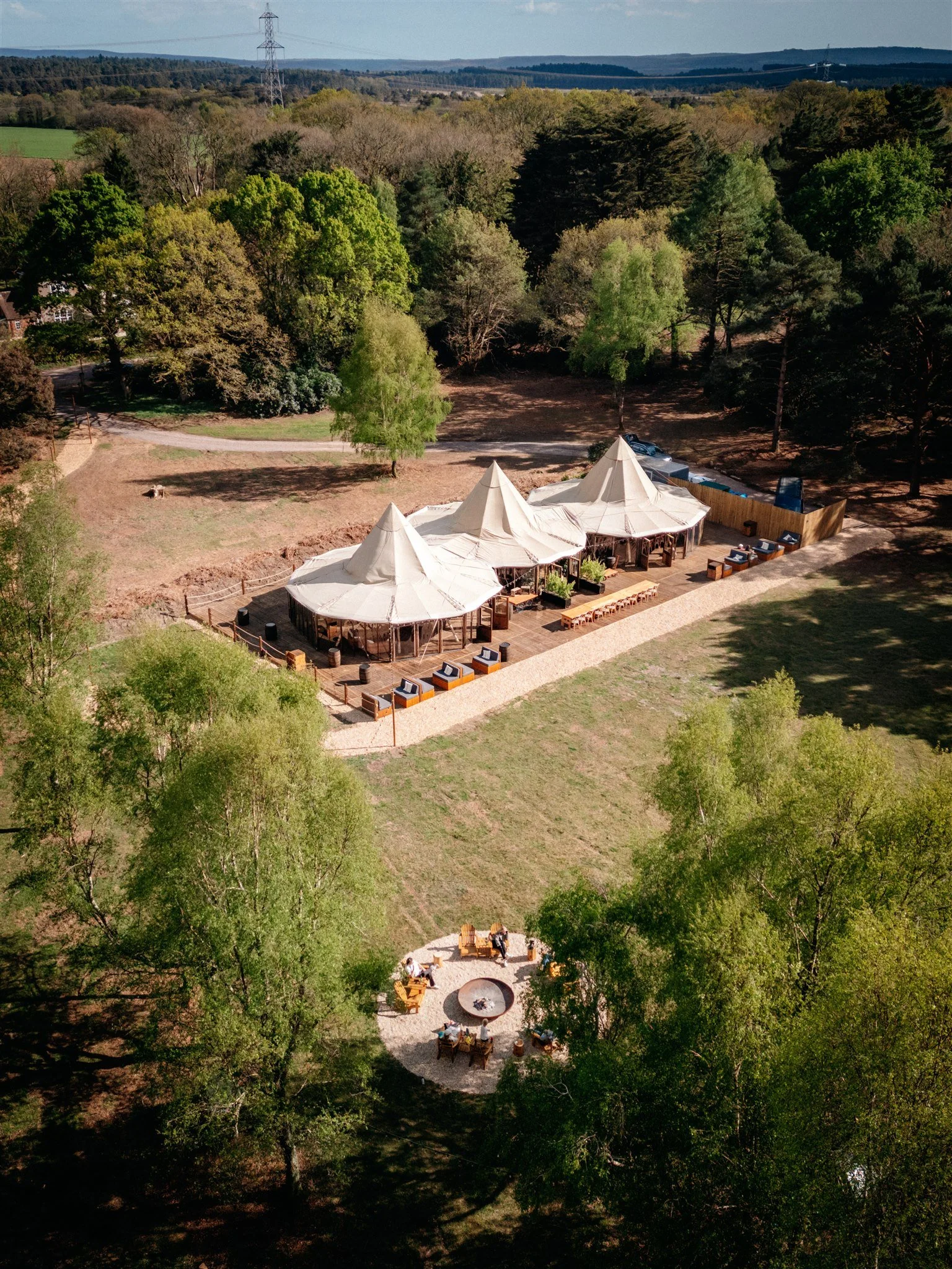 An aerial view of a large outdoor event setup in a park surrounded by trees, with multiple white tent structures, seating areas, and a fire pit, with a backdrop of rolling hills and power lines.