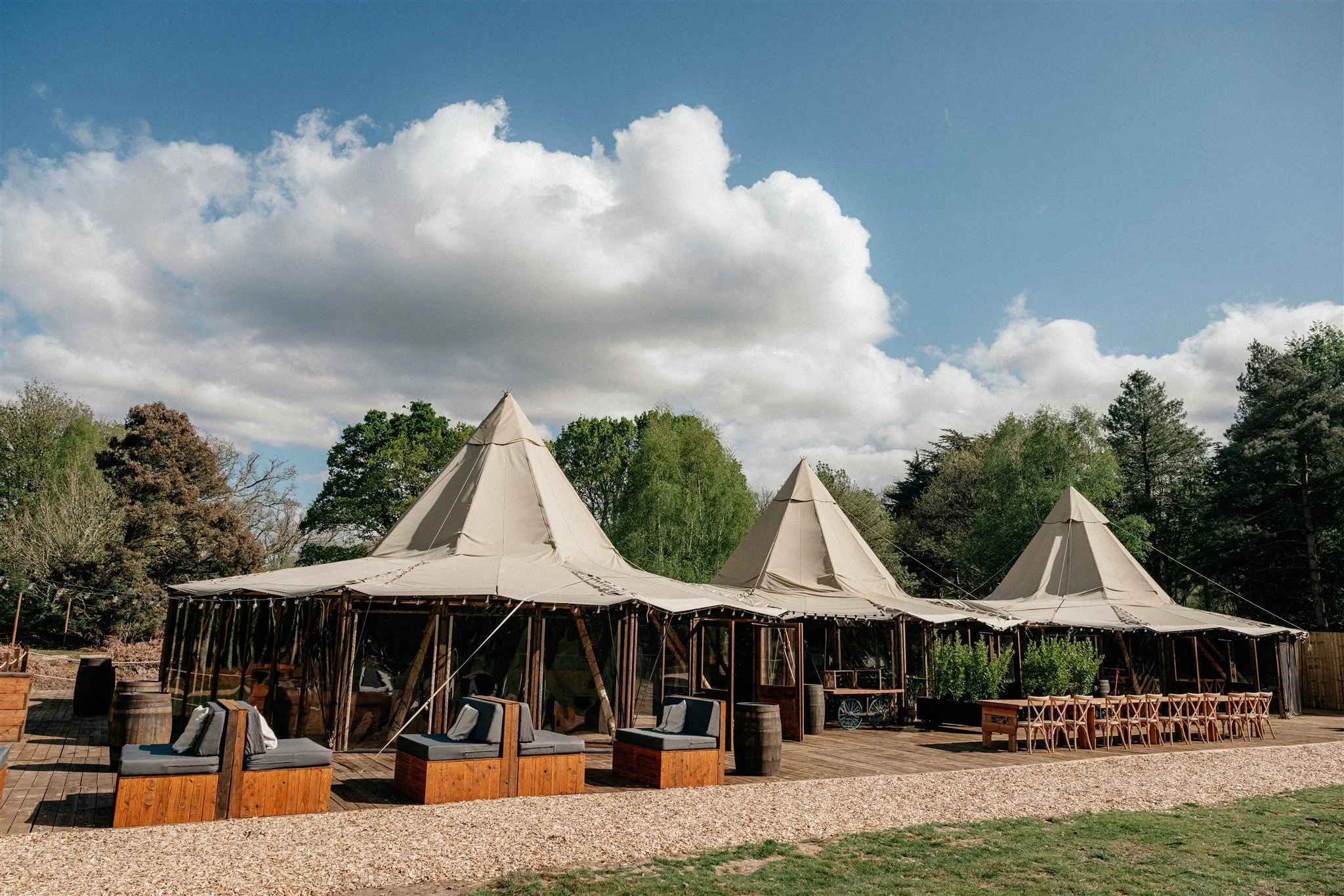 Arranged outdoor event space with several beige tipi tents, wooden tables, chairs, and lounge seating, surrounded by trees and under a partly cloudy sky.
