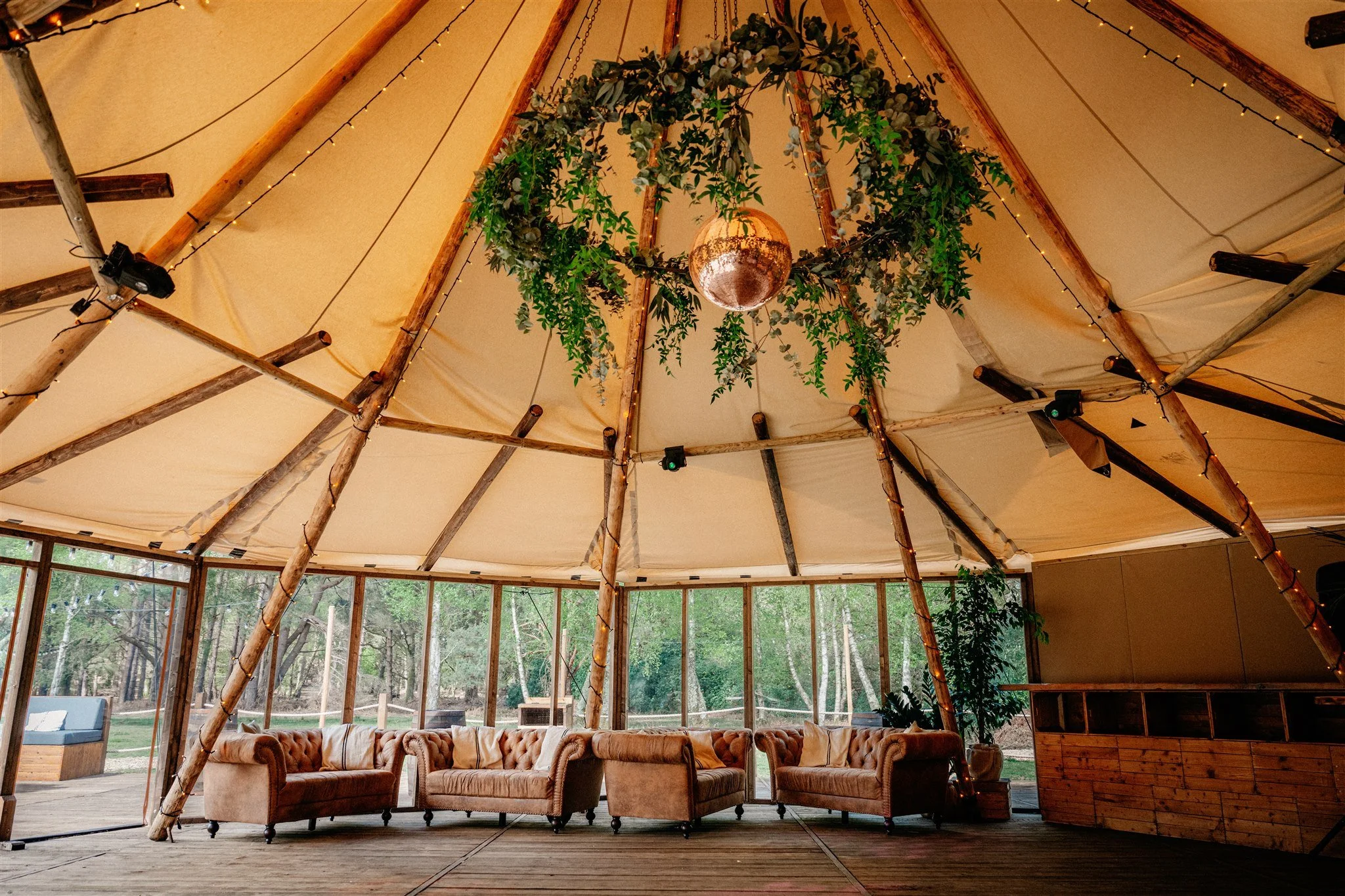 Interior of a tent with wooden supports, string lights, and a central chandelier with greenery. There are couches and a view of trees outside through glass windows.