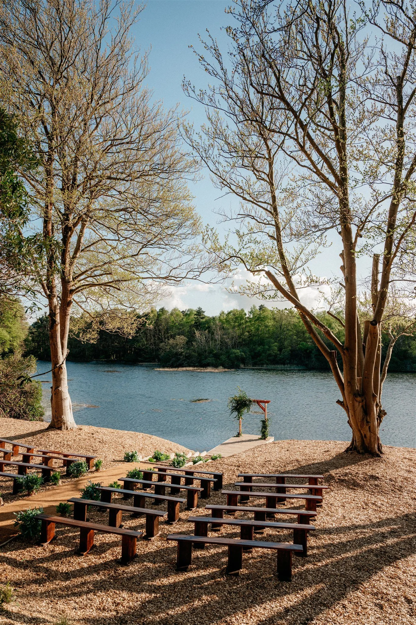 Outdoor wedding ceremony setup with wooden benches on gravel, a floral arch on a concrete platform near a river, and tall trees with budding leaves under a clear sky.