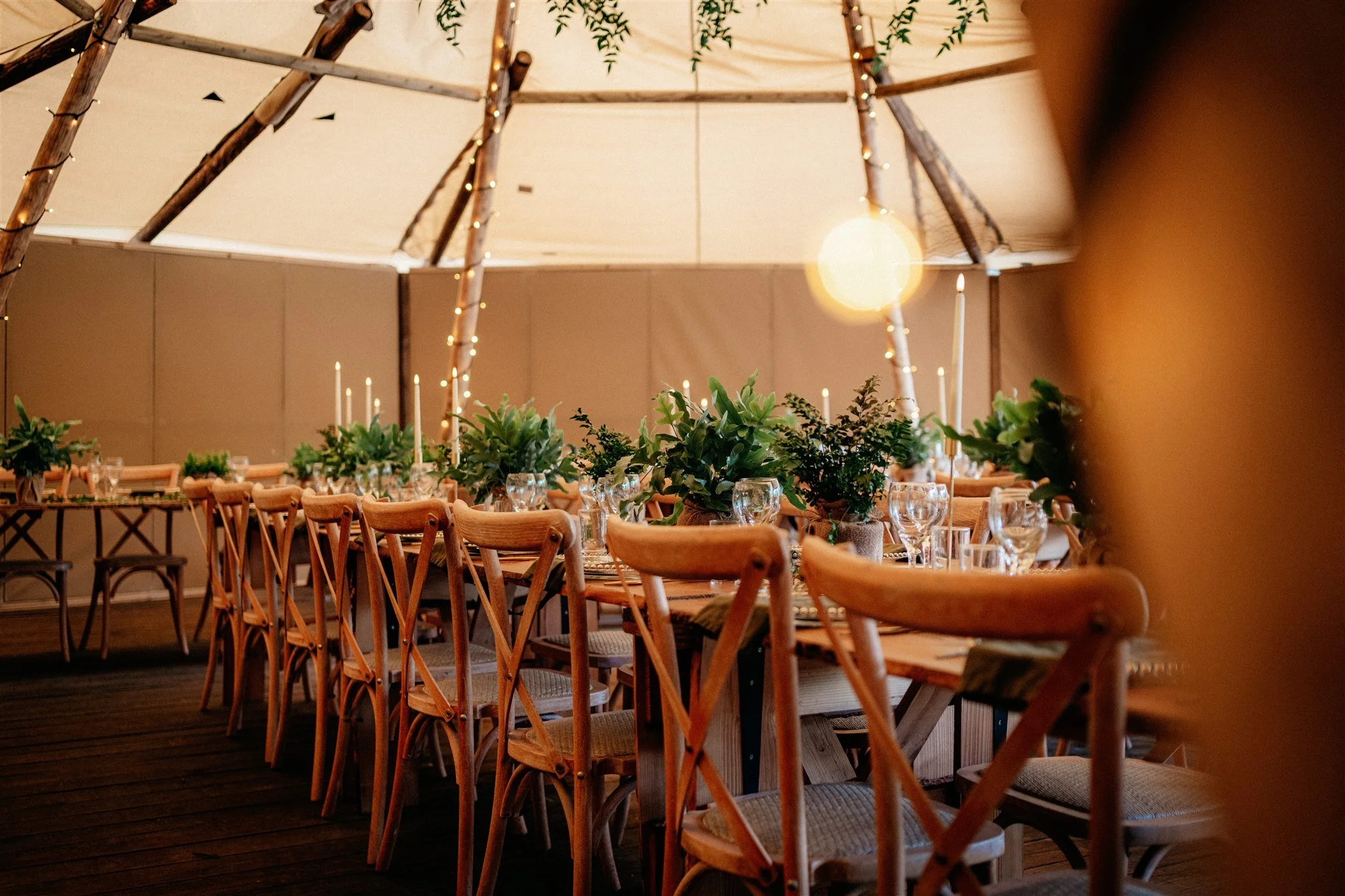 Decorative event table setting under a wooden and string-light structure with greenery and candle arrangements.