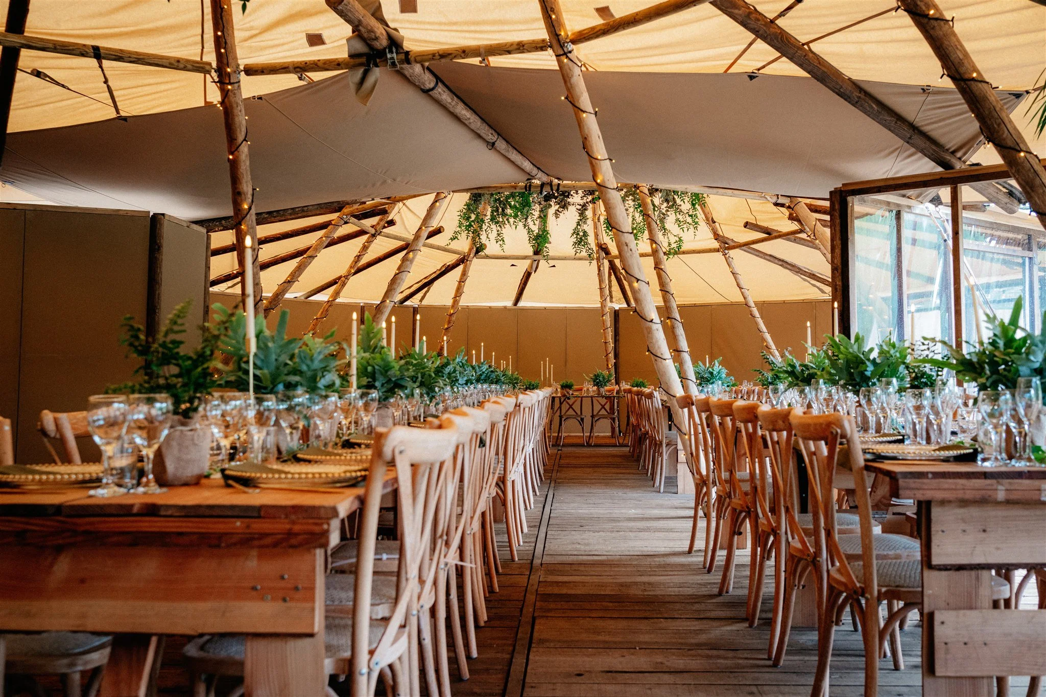 Inside a tent at an event, with a long dining table set with glassware, napkins, and greenery, surrounded by wooden chairs. The tent has a high ceiling with wooden beams, string lights, and hanging greenery.