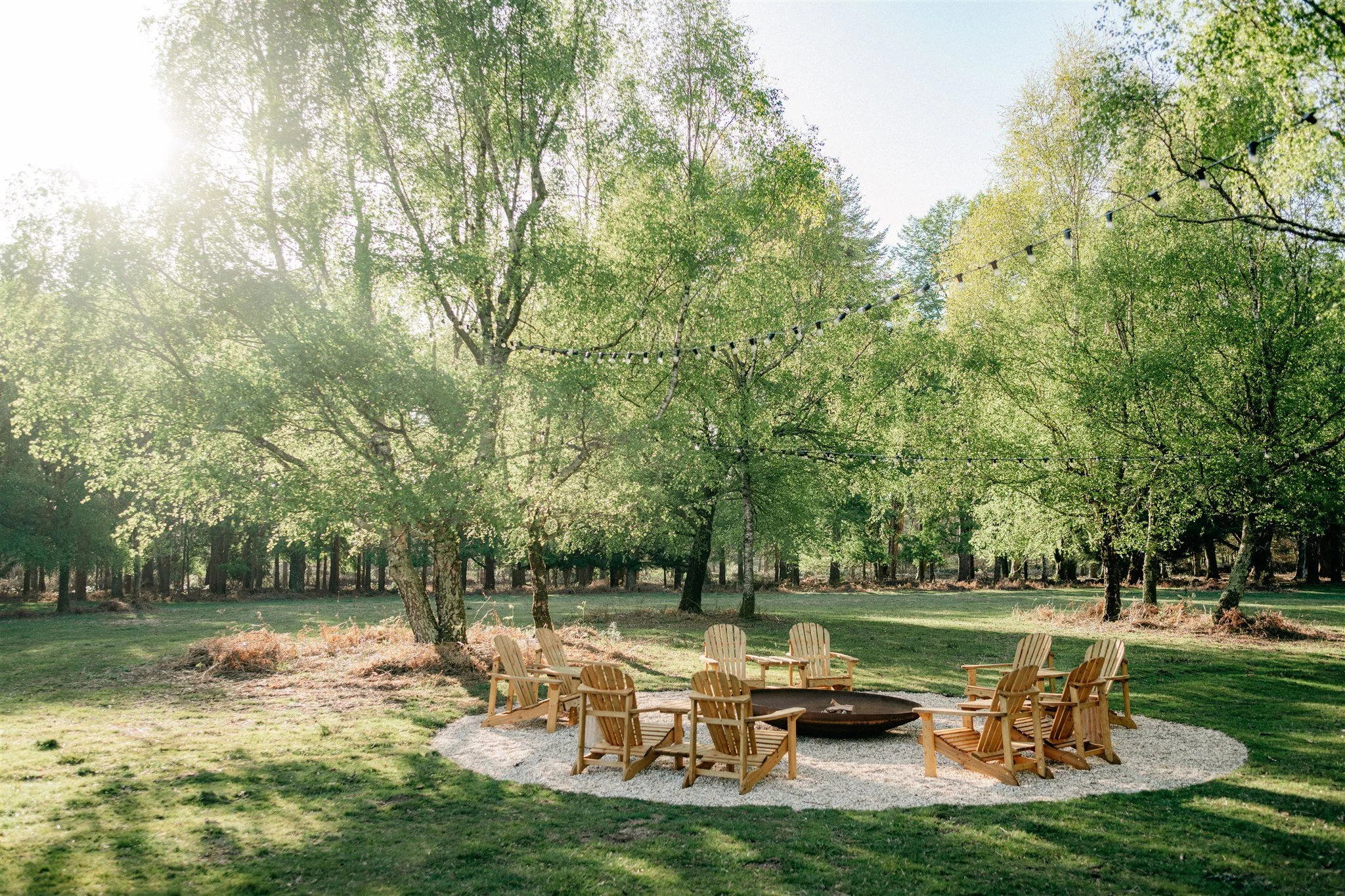 Outdoor gathering space with wooden chairs arranged around a fire pit, surrounded by green trees and string lights in a park setting on a sunny day.