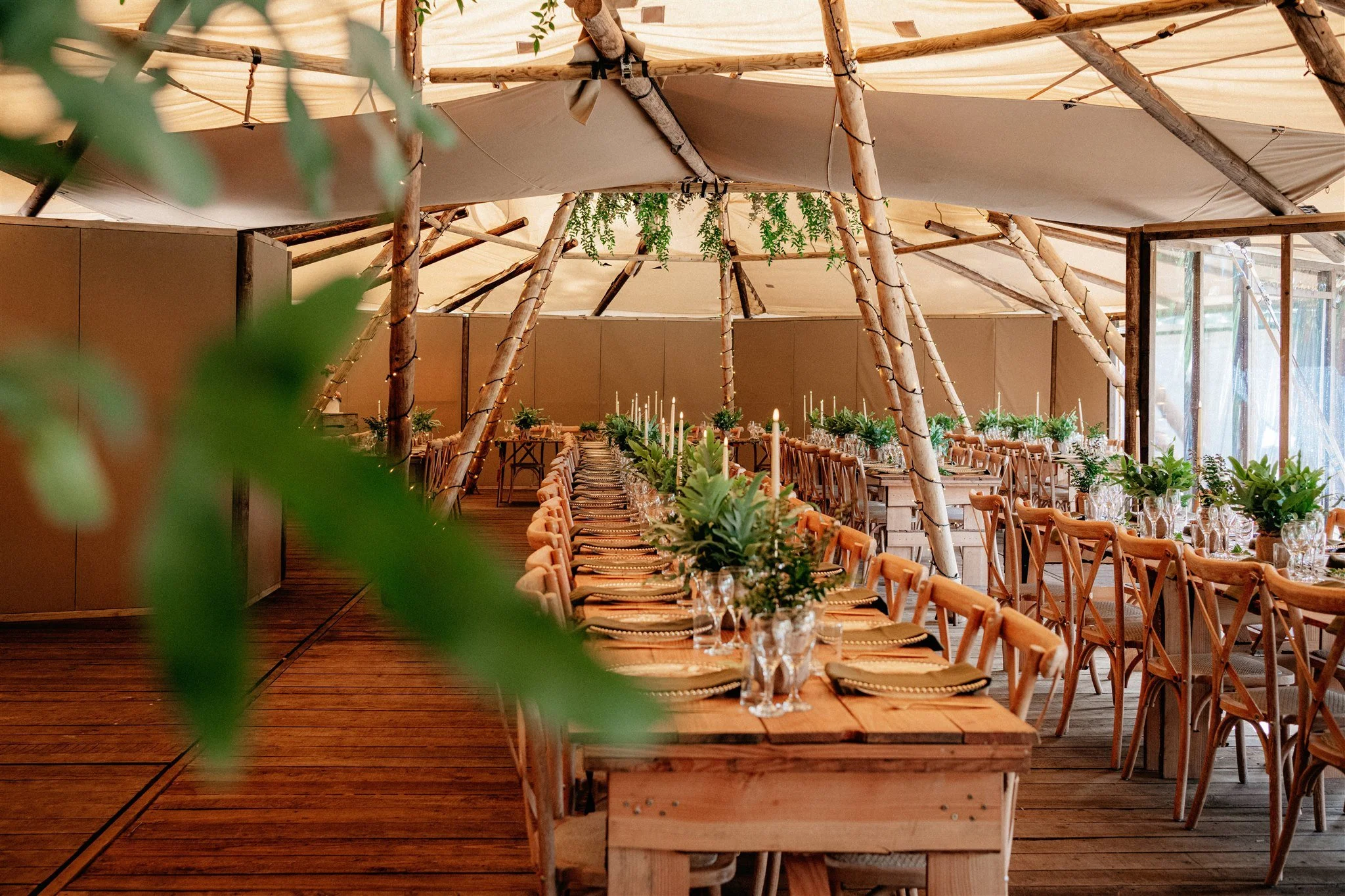 A rustic indoor event space with wooden furniture, long dining tables decorated with greenery and candles, and a tent-like ceiling with wooden beams and hanging vines.