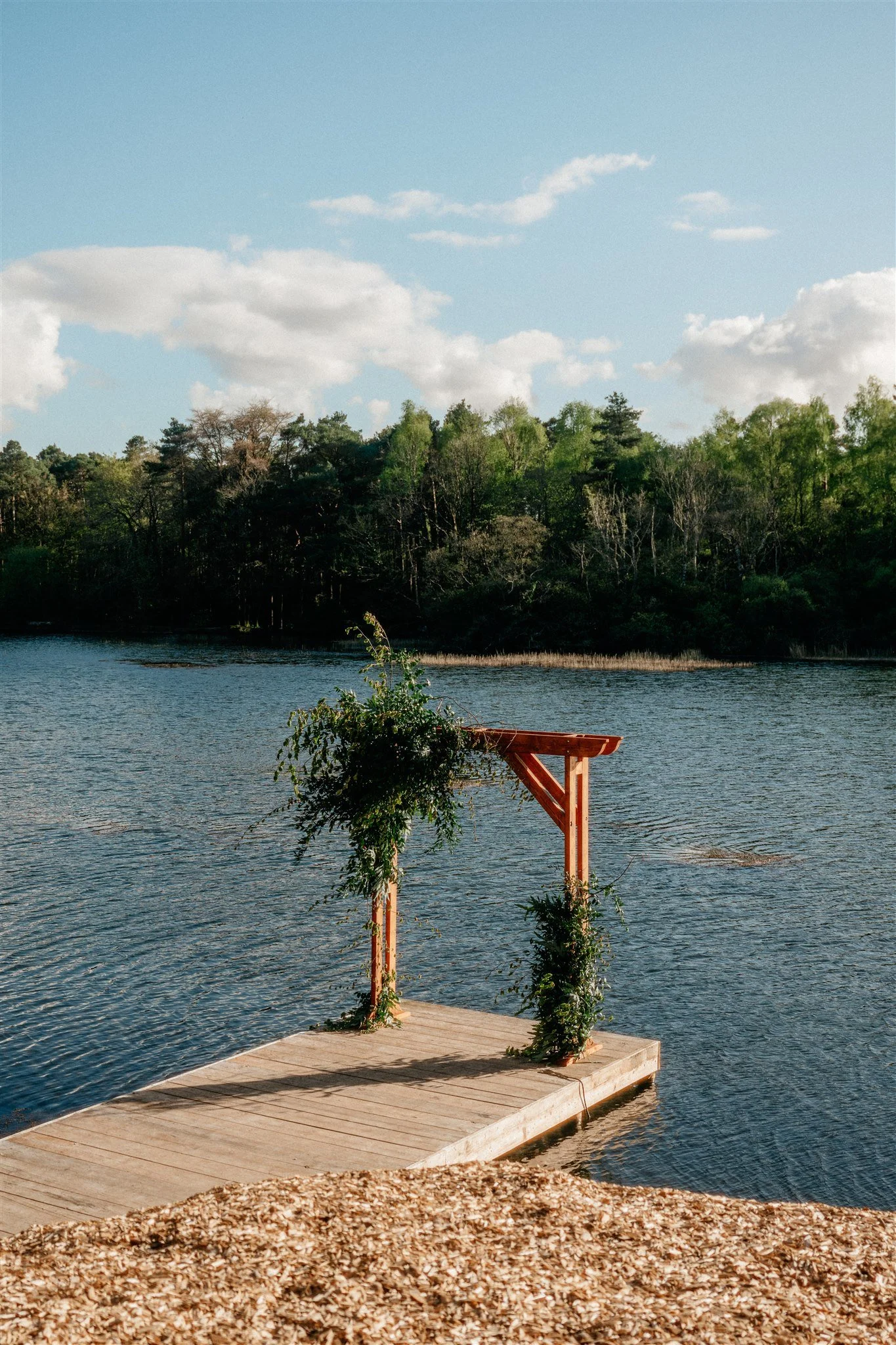 Lake with wooden dock and a small wooden structure with green vines, surrounded by trees and a partly cloudy sky.