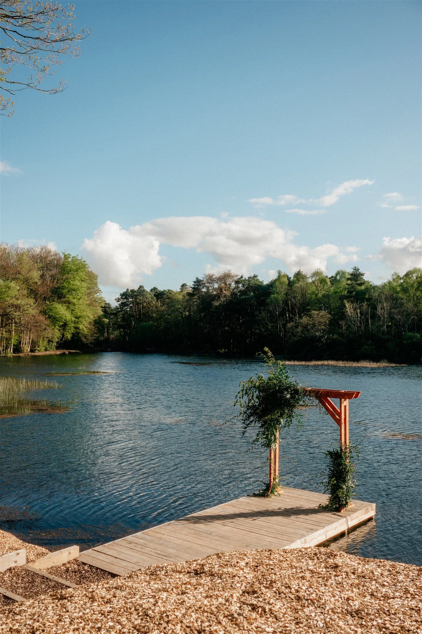 A lakeside scene with clear blue sky, trees along the shoreline, and a wooden dock with a small pergola decorated with vines extending over the water.