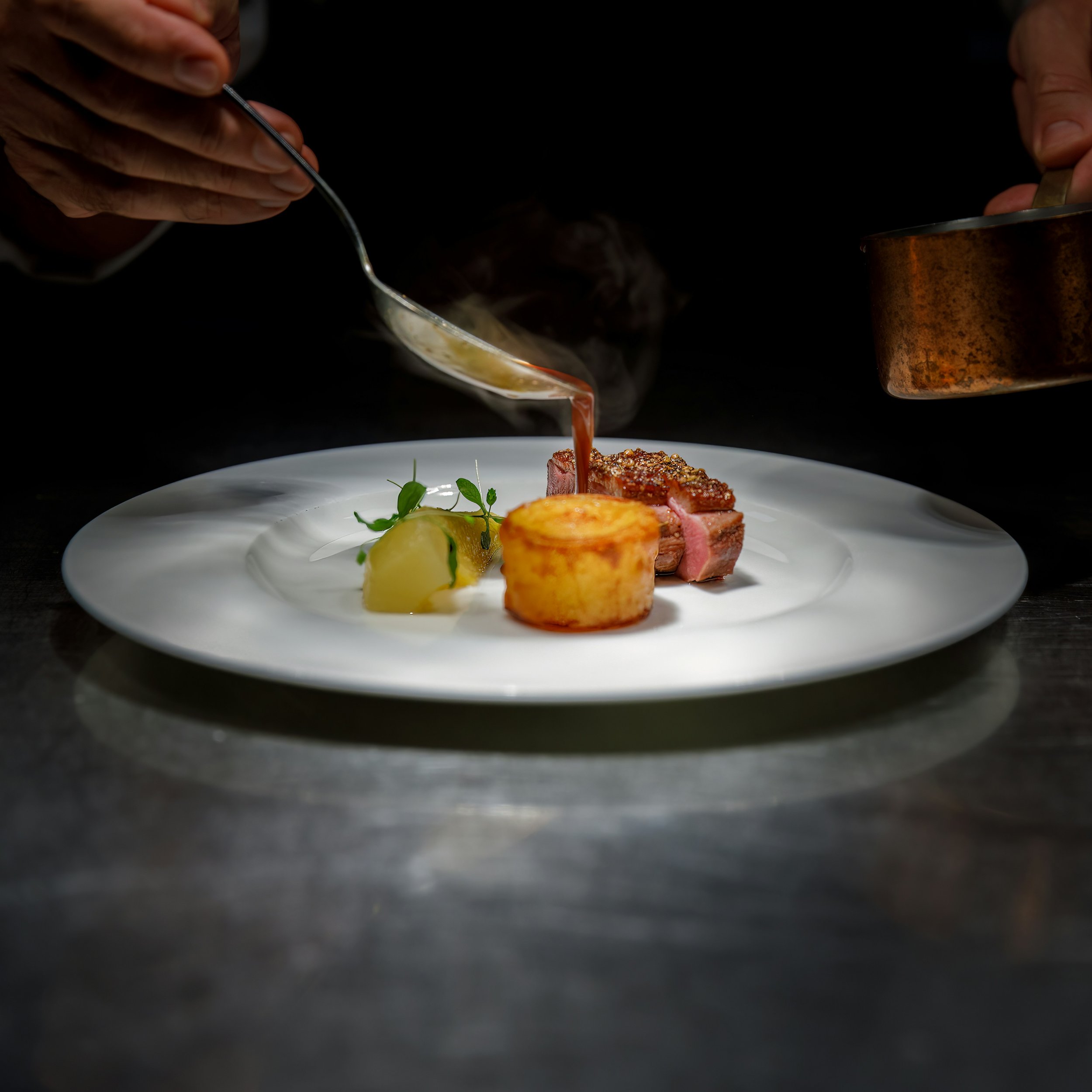 Chef pouring sauce over a plated dish with sliced steak, a roasted vegetable, and garnished with herbs on a white plate.