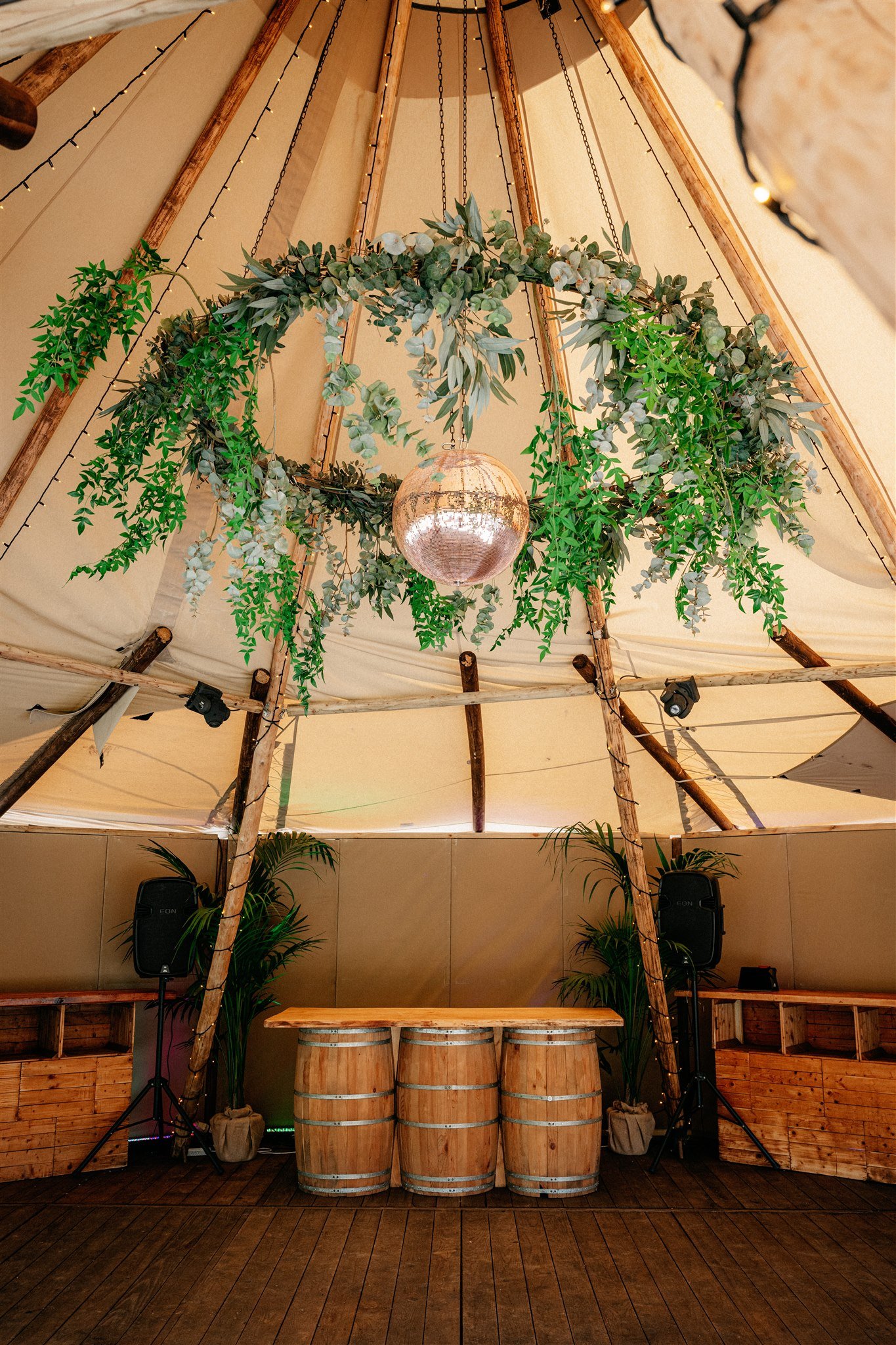 Interior of a tent-style event space decorated with green leafy garlands hanging from wooden beams and a disco ball at the center. There are speakers, potted plants, and wooden furniture including barrels and benches.