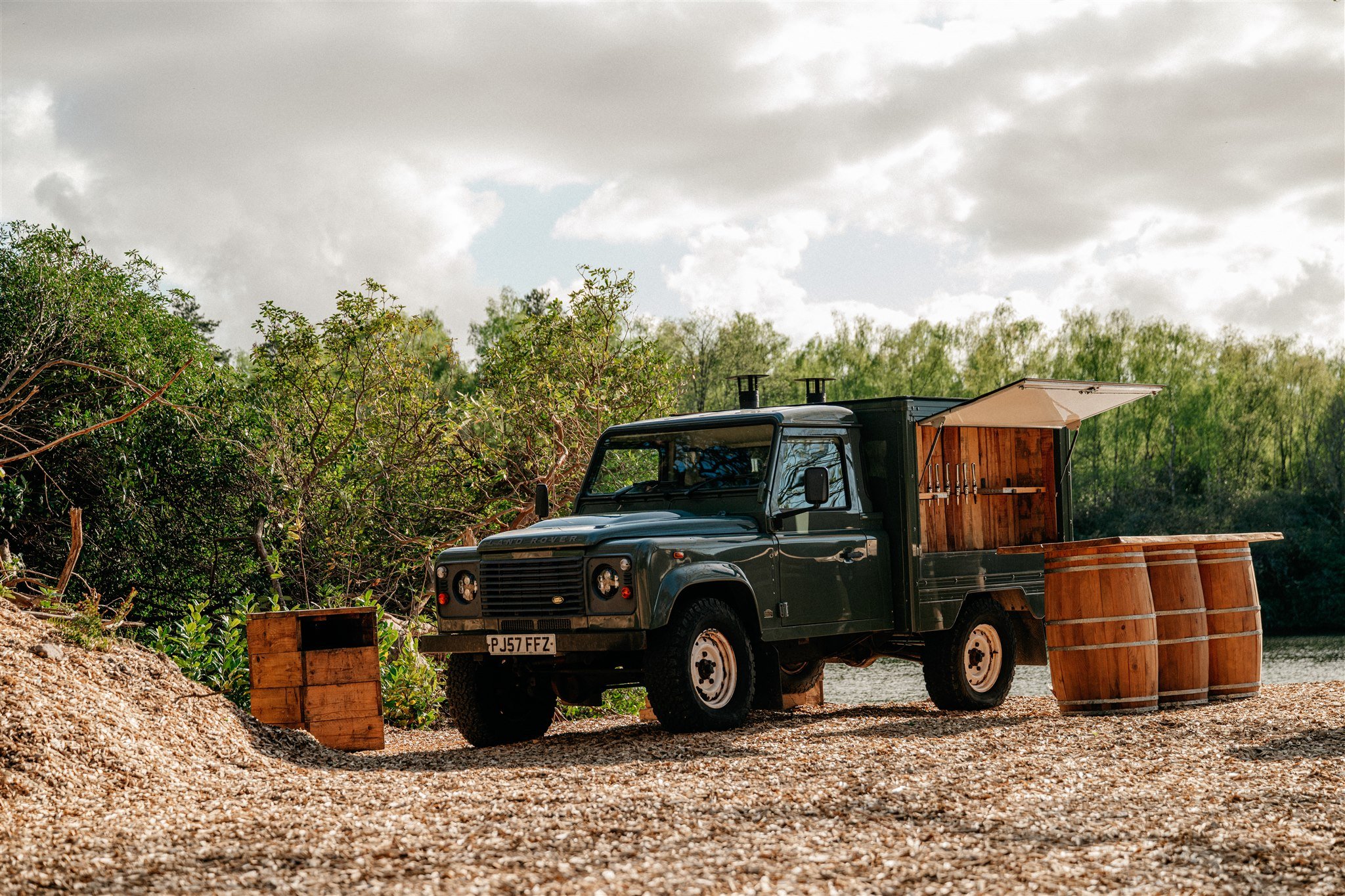 A black Land Rover Defender parked outdoors next to a large pile of gravel and wooden barrels, with lush green trees and a cloudy sky in the background.