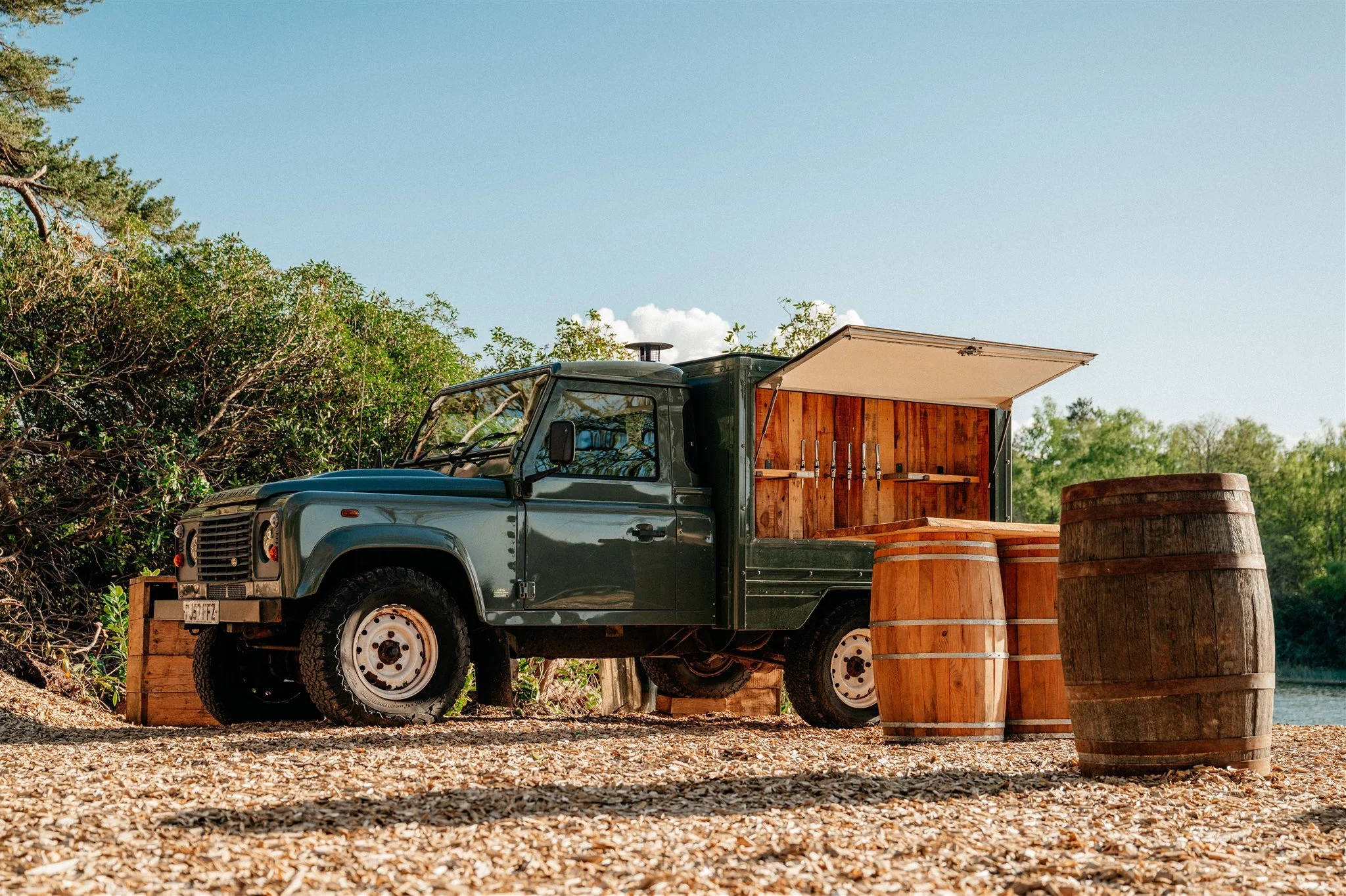 A dark green vintage off-road vehicle with a wooden side panel is parked on a gravel surface. Large wooden barrels are placed nearby, and the vehicle is elevated on wooden blocks. Surrounding trees and bushes are in the background under a clear blue 