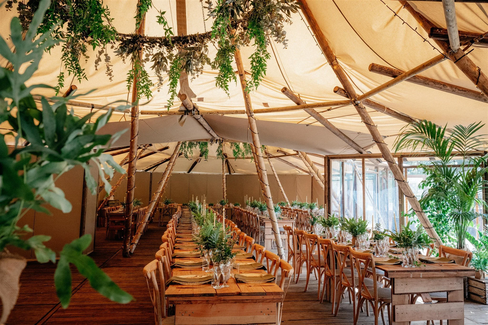 Inside a rustic wedding reception venue decorated with bamboo and greenery, with arranged tables, chairs, and centerpieces, under a canopy of beige fabric and string lights.