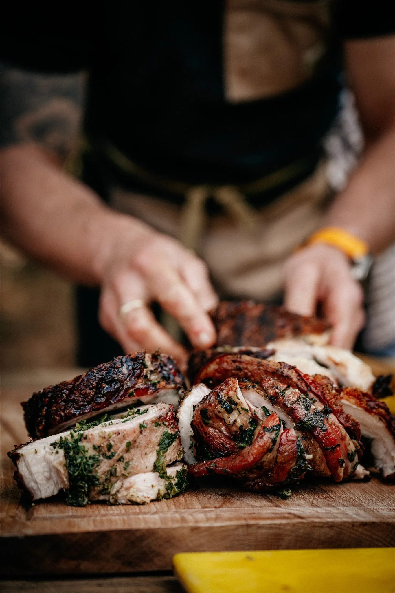 Close-up of sliced grilled meat with herbs on a wooden cutting board, with person in the background preparing or serving it.