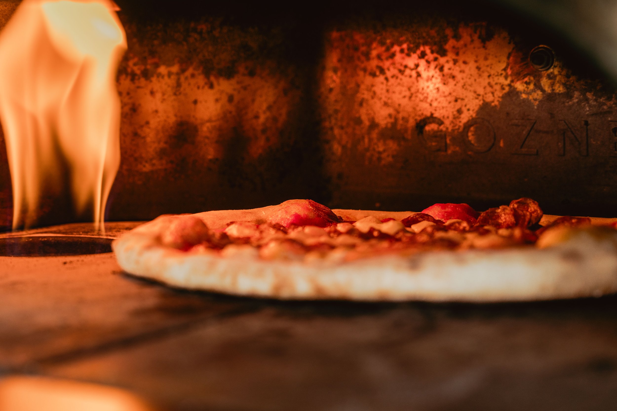 Pizza baking inside a wood-fired oven with flames visible on the left side.