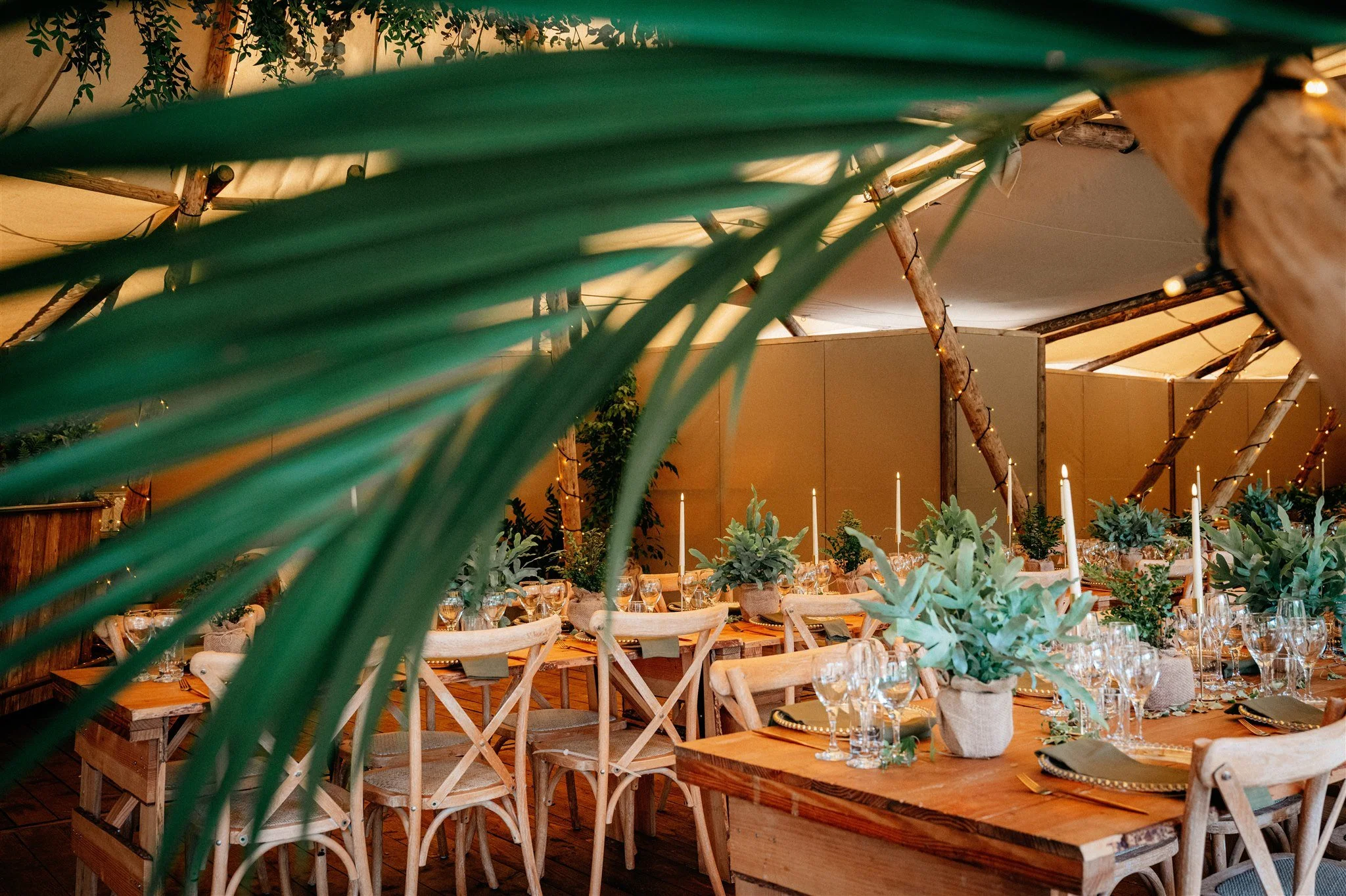 A rustic indoor dining area decorated with green plants, string lights, and candles on the tables, with wooden tables and chairs arranged for a gathering.