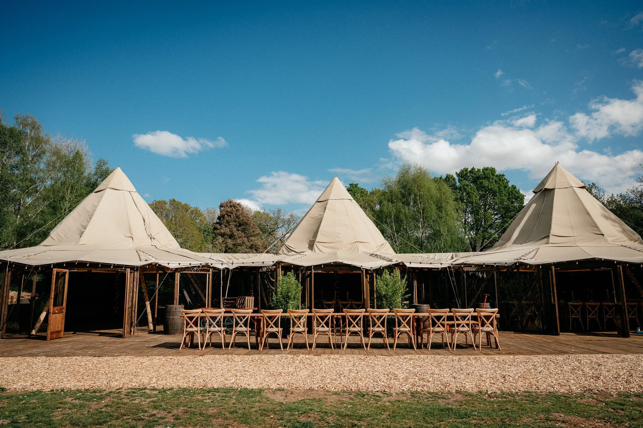 Outdoor scene with a large beige tent structure made of fabric and wooden support beams, surrounded by trees under a blue sky with scattered clouds.