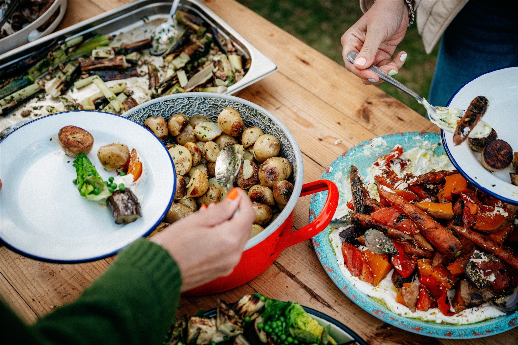A variety of grilled vegetables, including zucchini, mushrooms, tomatoes, and eggplant, are displayed on large plates and trays on a wooden table at an outdoor gathering, with a person serving food.