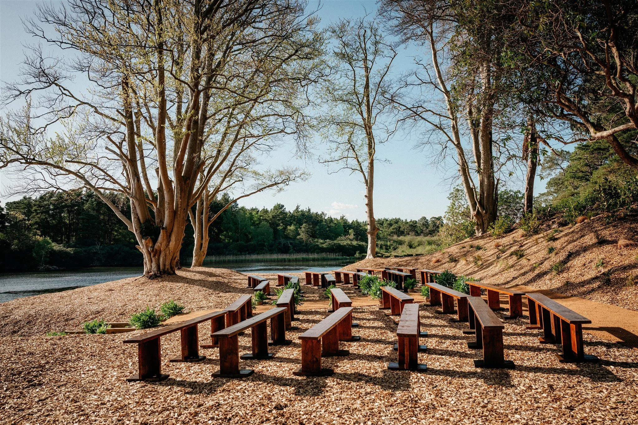 Wooden benches arranged in rows on a gravel area by a river, with tall trees on a hillside and a clear blue sky in the background.