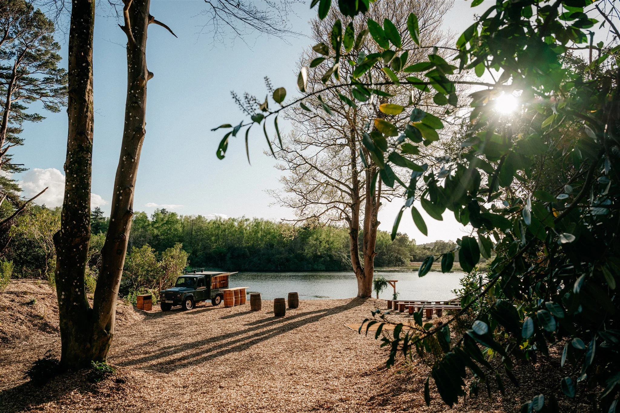 View of a lakeside with trees, a boat, and chairs, sunlight filtering through foliage.