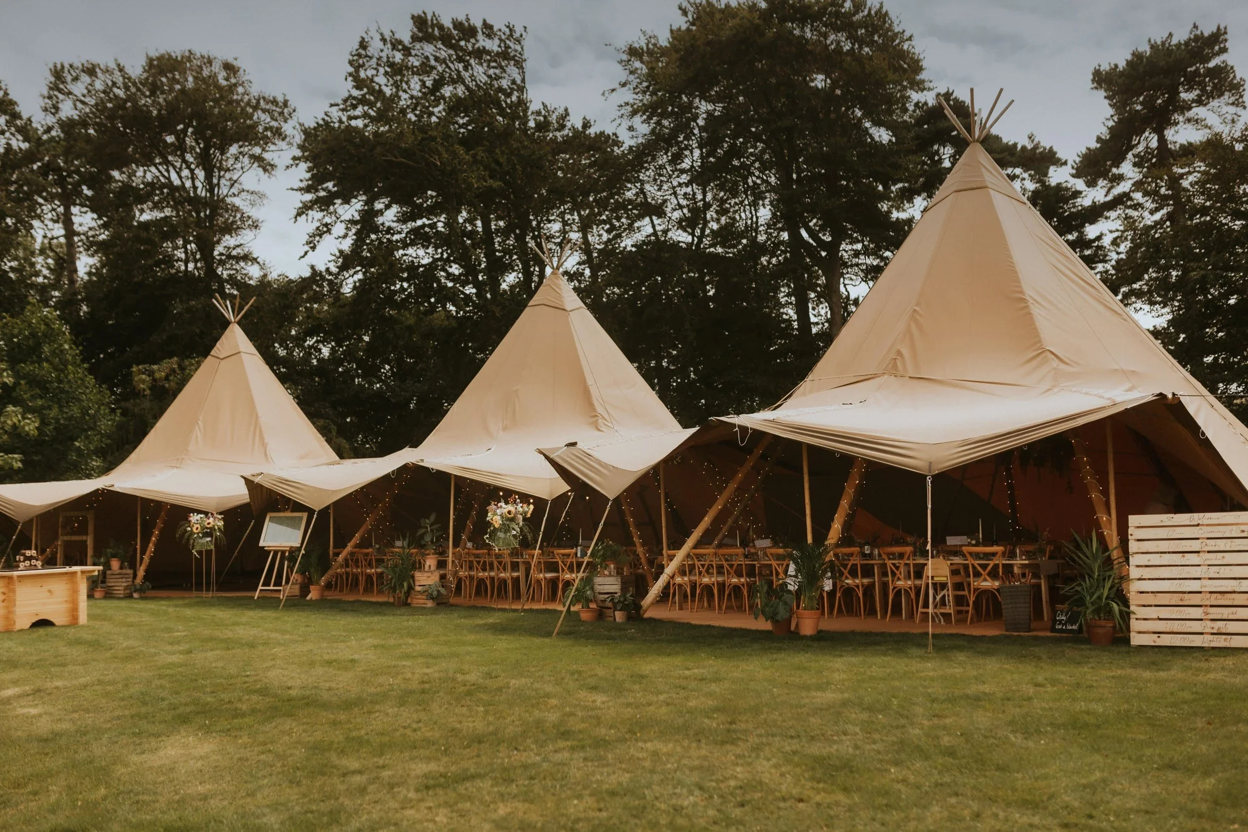 A large outdoor event tent with a teepee style design, set up on a grassy area with trees in the background. The tent has string lights and plants around it, with wooden chairs and tables inside, likely for a celebration or gathering.