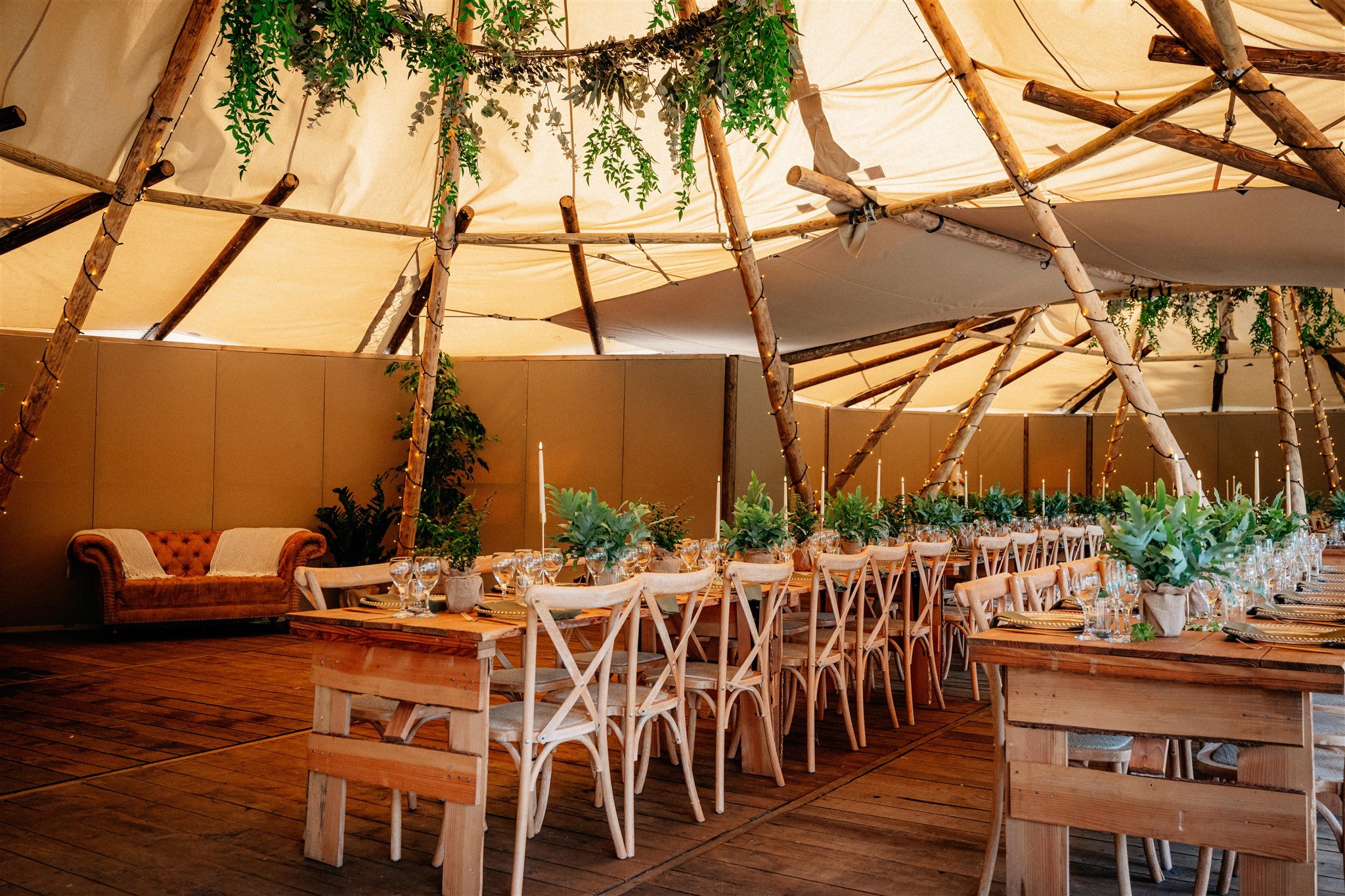 Indoor event space decorated with wooden furniture, green plants, string lights, and draped fabric ceiling, set up for a gathering or celebration.