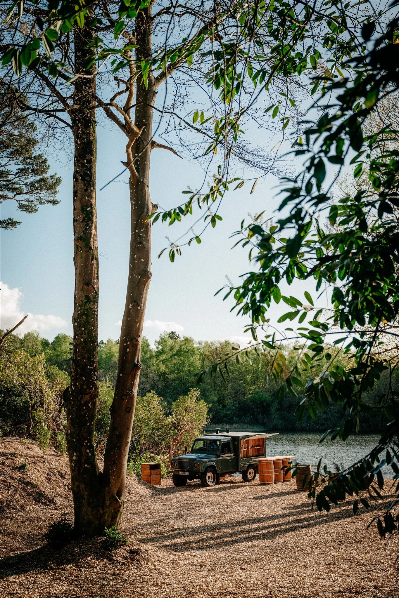 A serene lakeside scene with trees and a boat, green foliage on the trees, and a vintage truck parked near the water with barrels nearby.