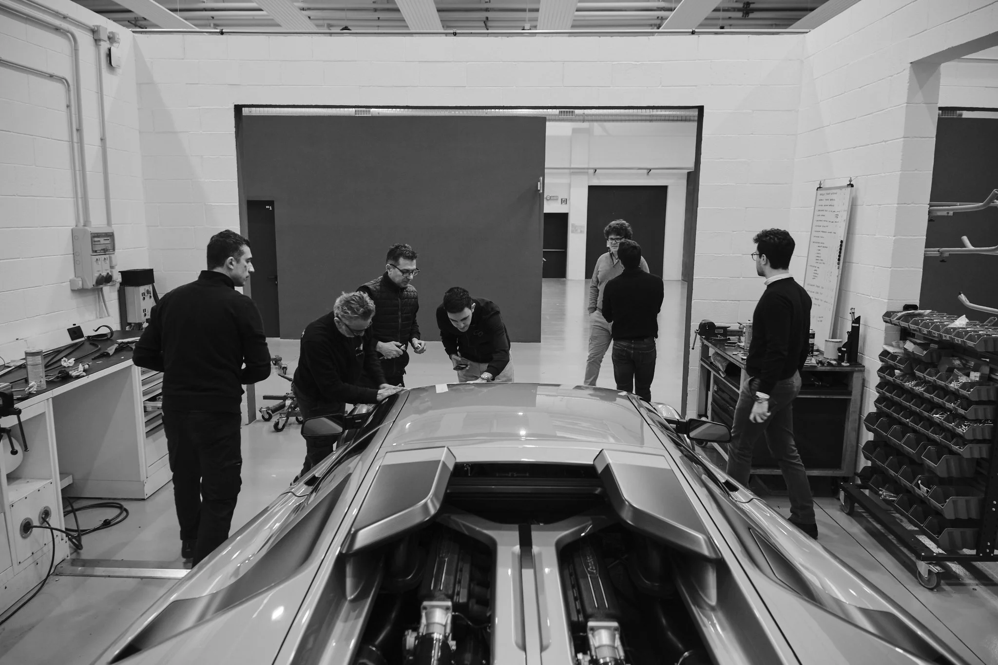 Group of people in a workshop or garage examining a sports car with an open engine hood, surrounded by tools and equipment.