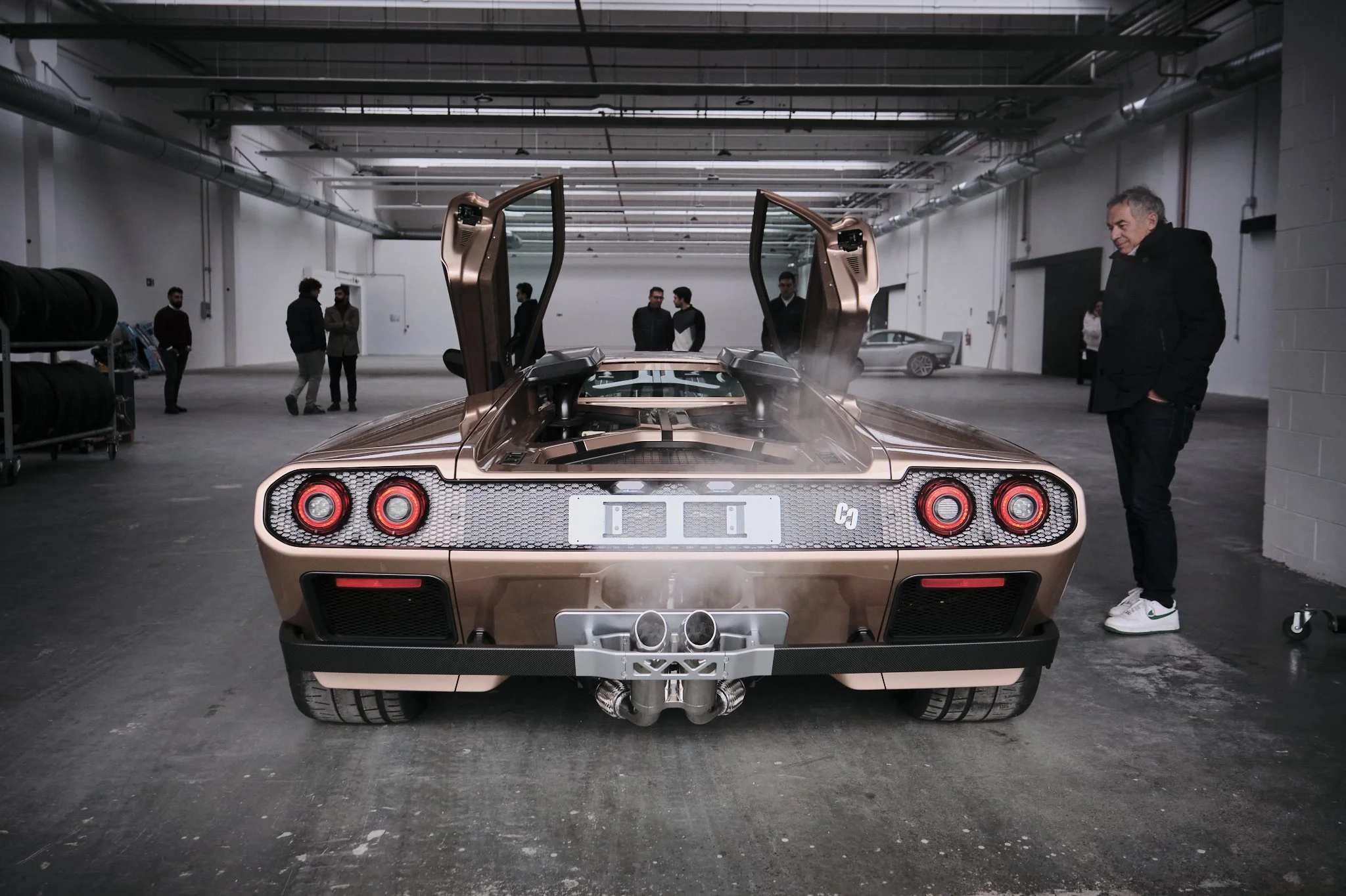 Rear view of a bronze-colored Lamborghini Countach with butterfly doors open, parked in an indoor garage with people observing.