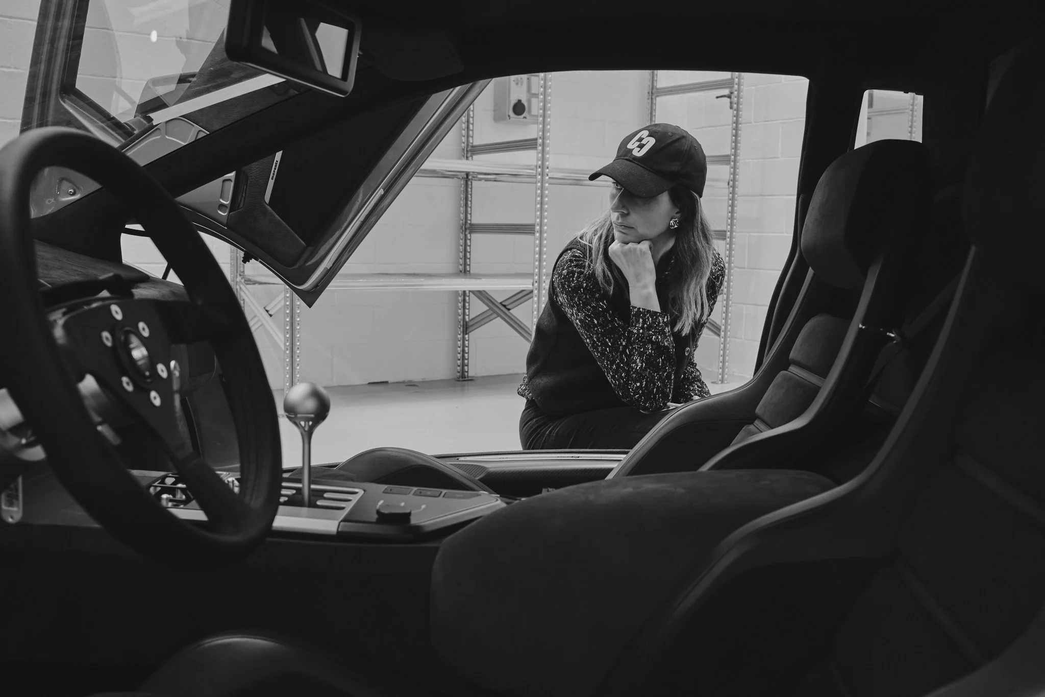 A woman sitting outside a vintage sports car, looking thoughtfully at the car's interior, in a garage setting.