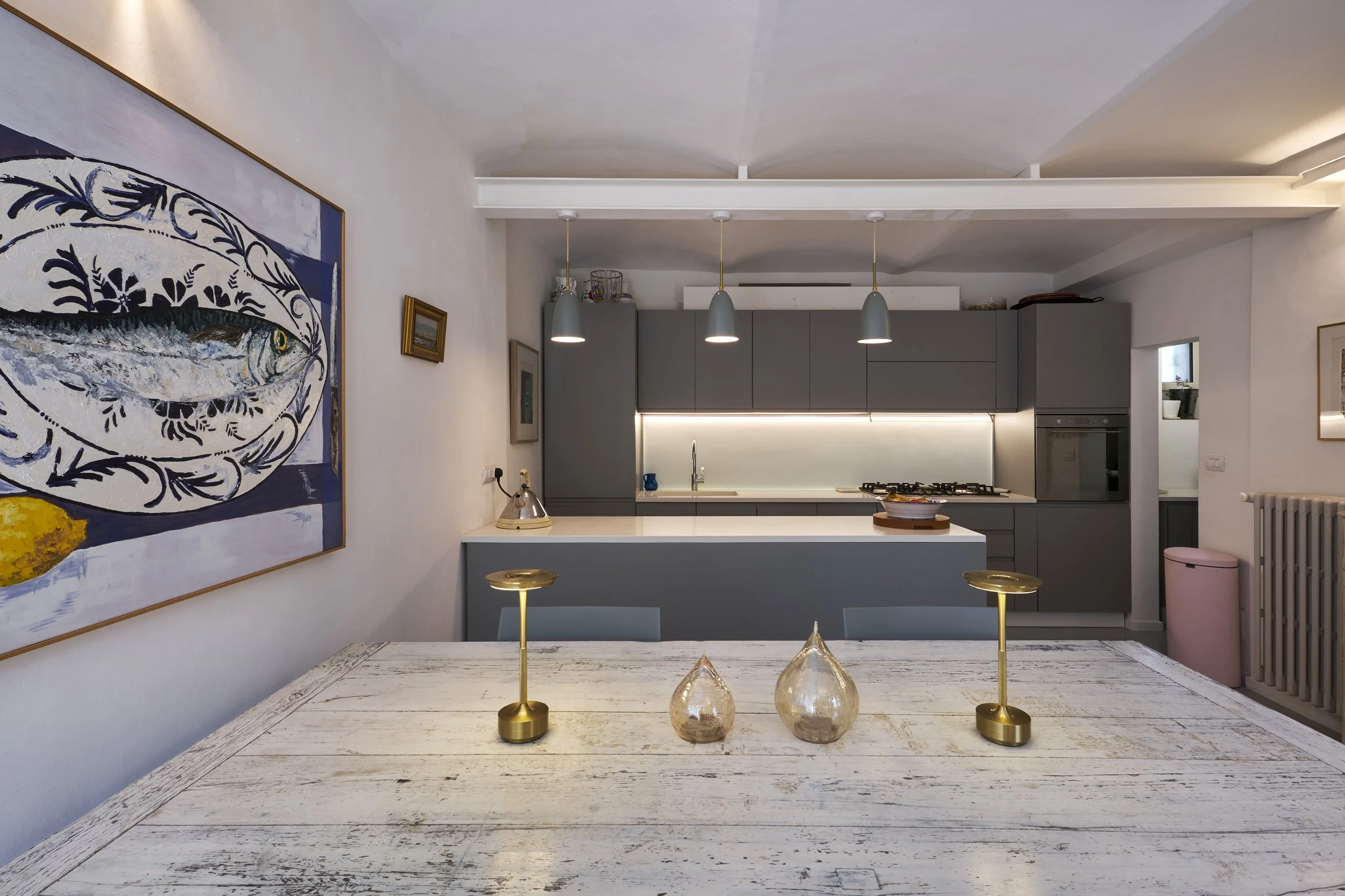 Modern kitchen with grey cabinetry, white countertop, hanging pendant lights, and a table with three decorative candles in glass holders, adjacent to a dining table with vintage white paint finish.