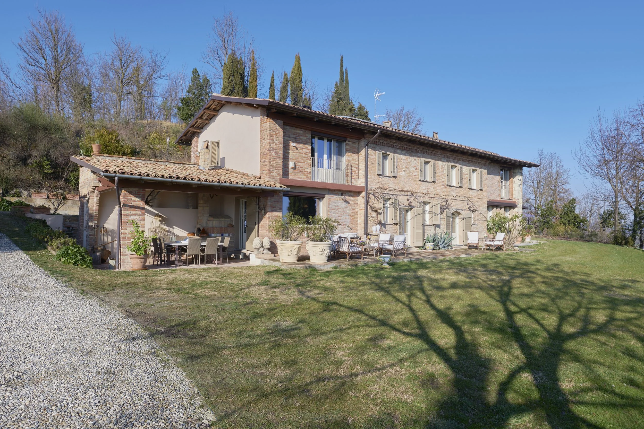 Large rustic house with brick and stucco exterior, surrounded by green lawn and patio furniture, with leafless trees and a clear blue sky in the background.
