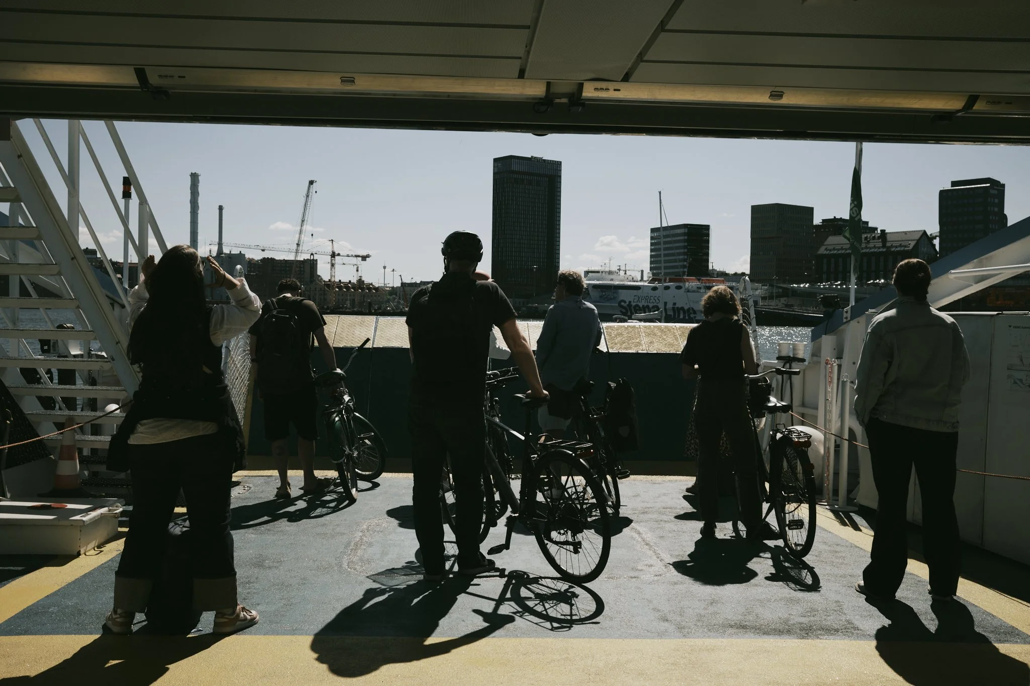 People waiting with bicycles on a ferry in a city harbor, with tall buildings and a boat in the background.