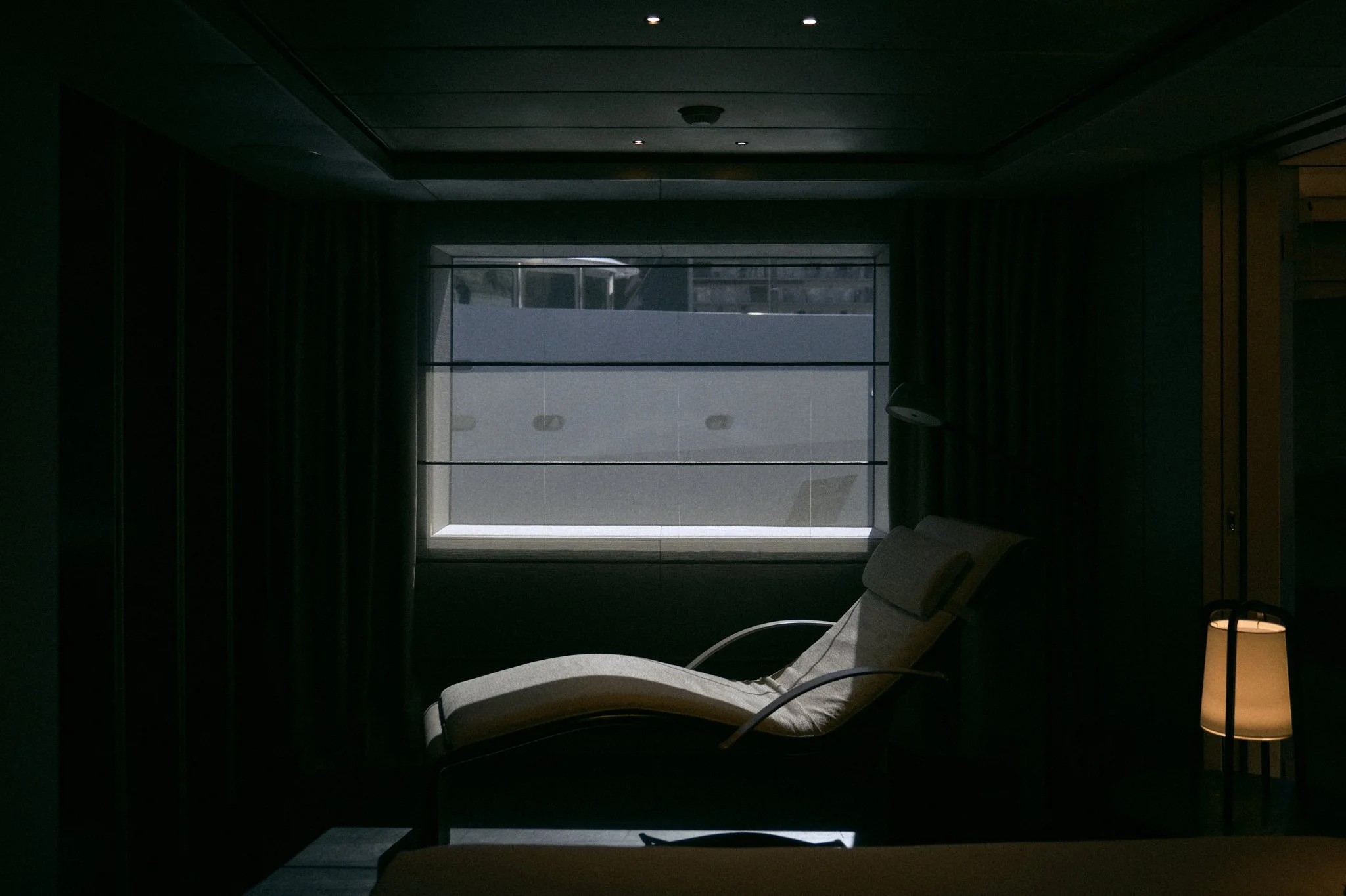 A dimly lit hotel room with a lounge chair near a window, and a standing lamp emitting warm light.