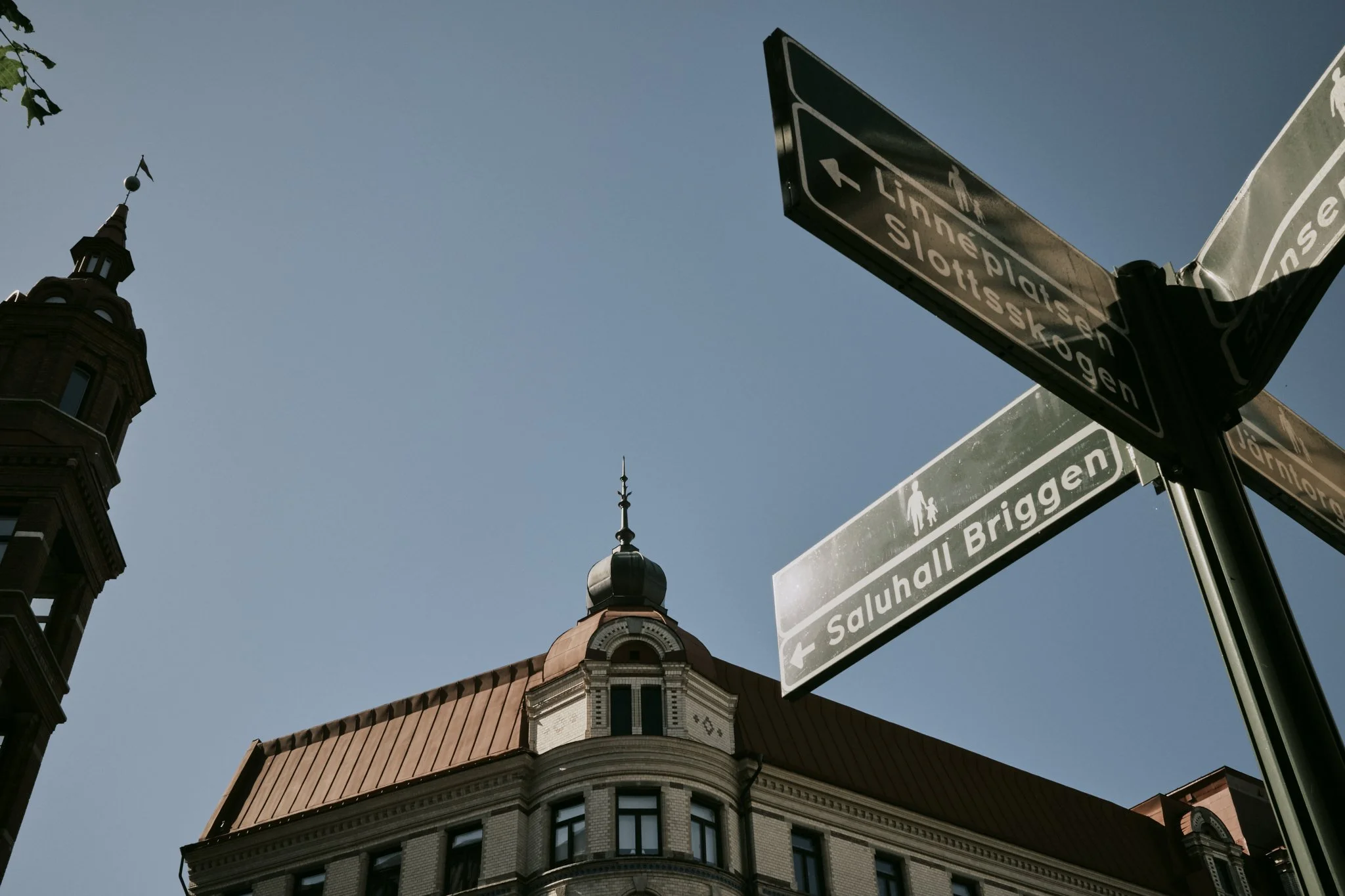 Street signs on a pole pointing to Tinerplatz, Slottskogen, and Saluhall Brggen, with a historical building with a red roof and tower in the background against a clear blue sky.