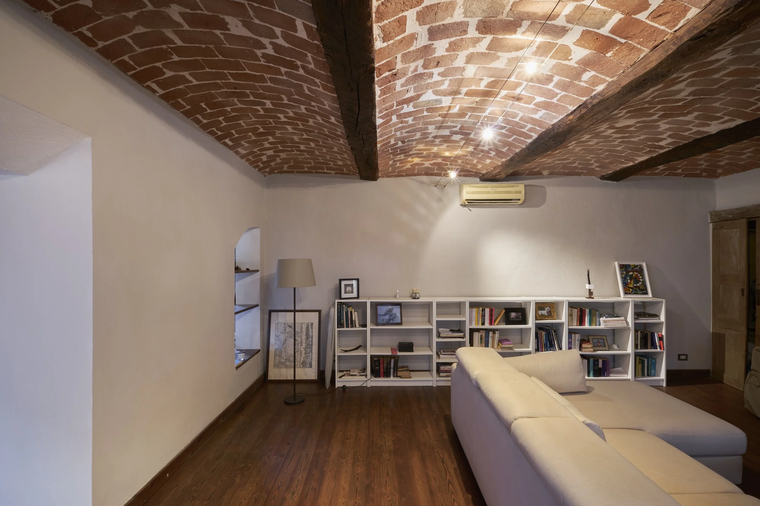 Living room with a brick vaulted ceiling, a white couch, a tall floor lamp, a white bookshelf with books and framed pictures, and wooden flooring.