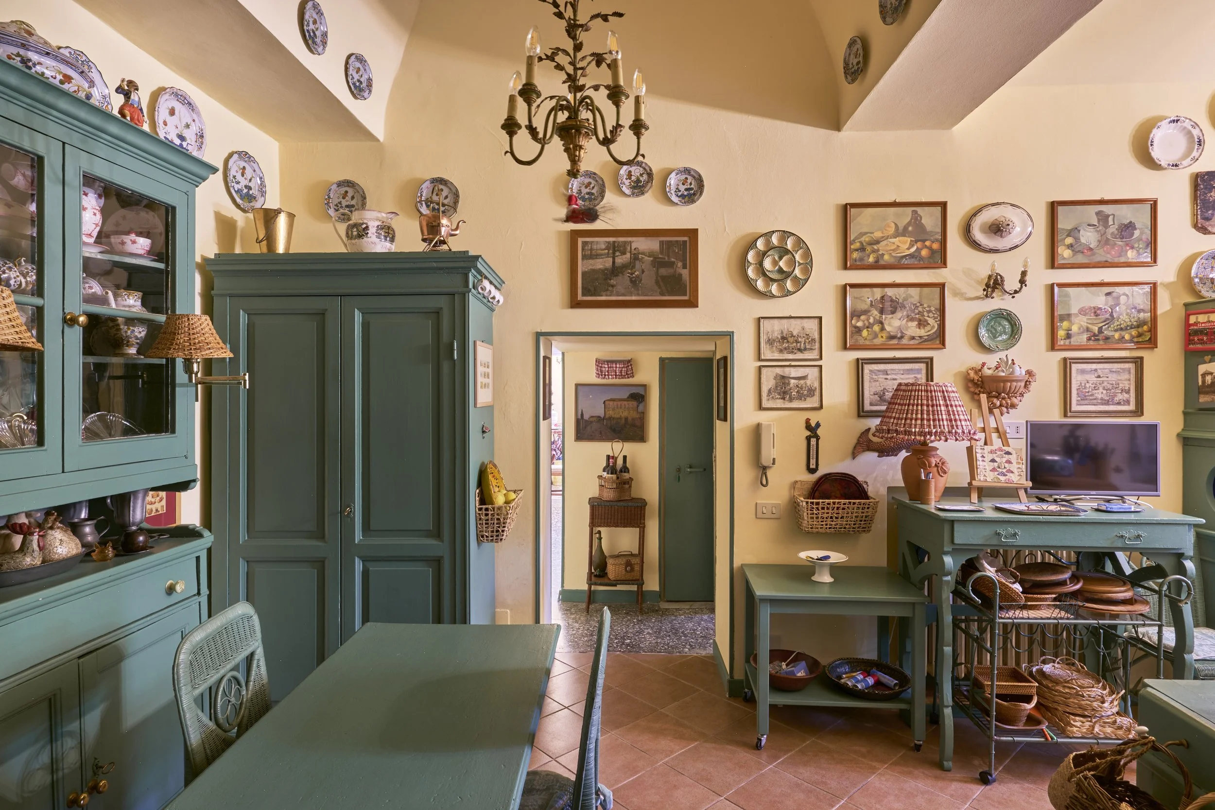 A cozy living room with yellow walls decorated with plates, framed pictures, and a chandelier. There are green cabinets, tables, and a small flat-screen TV. Wicker baskets and vintage decor fill the space.