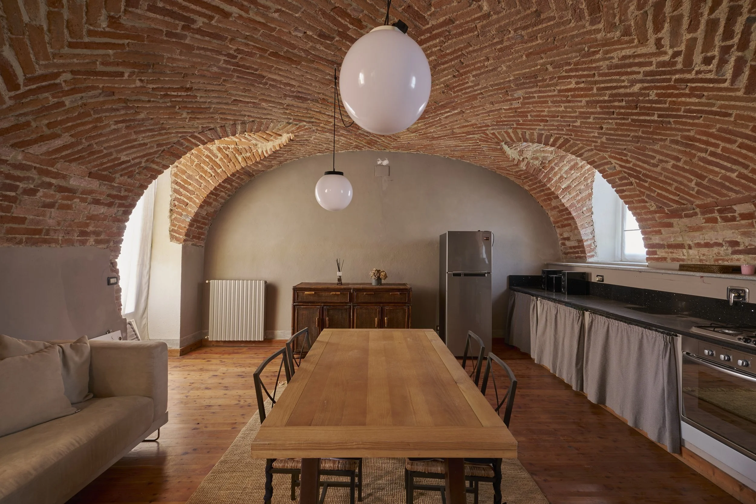 Interior of a rustic kitchen with a vaulted brick ceiling, wooden floor, a wooden dining table with six black chairs, a beige sofa, a gray refrigerator, a wooden sideboard, and a long black countertop with a curtain underneath, with windows providing