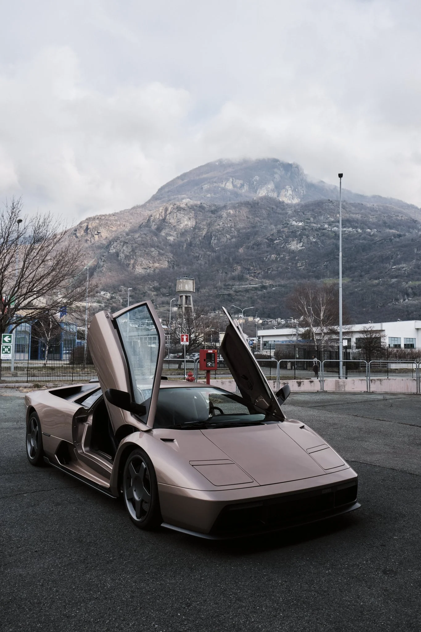 A beige Lamborghini Countach with scissor doors open parked in an outdoor lot with a mountain in the background.