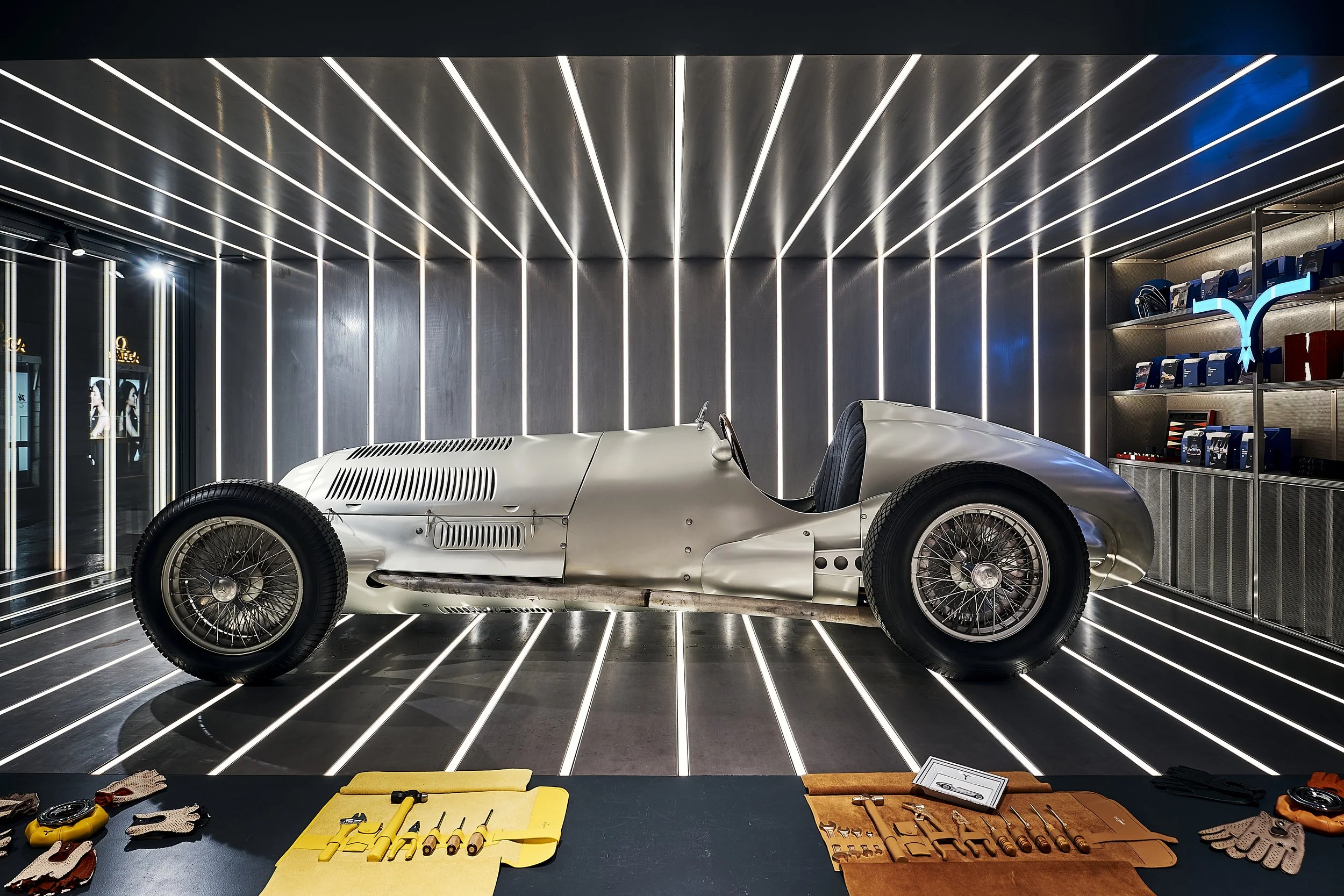 Vintage silver racing car inside a modern showroom with illuminated black walls, tools and gloves on a black surface in the foreground, and a display shelf with helmets and items on the right.