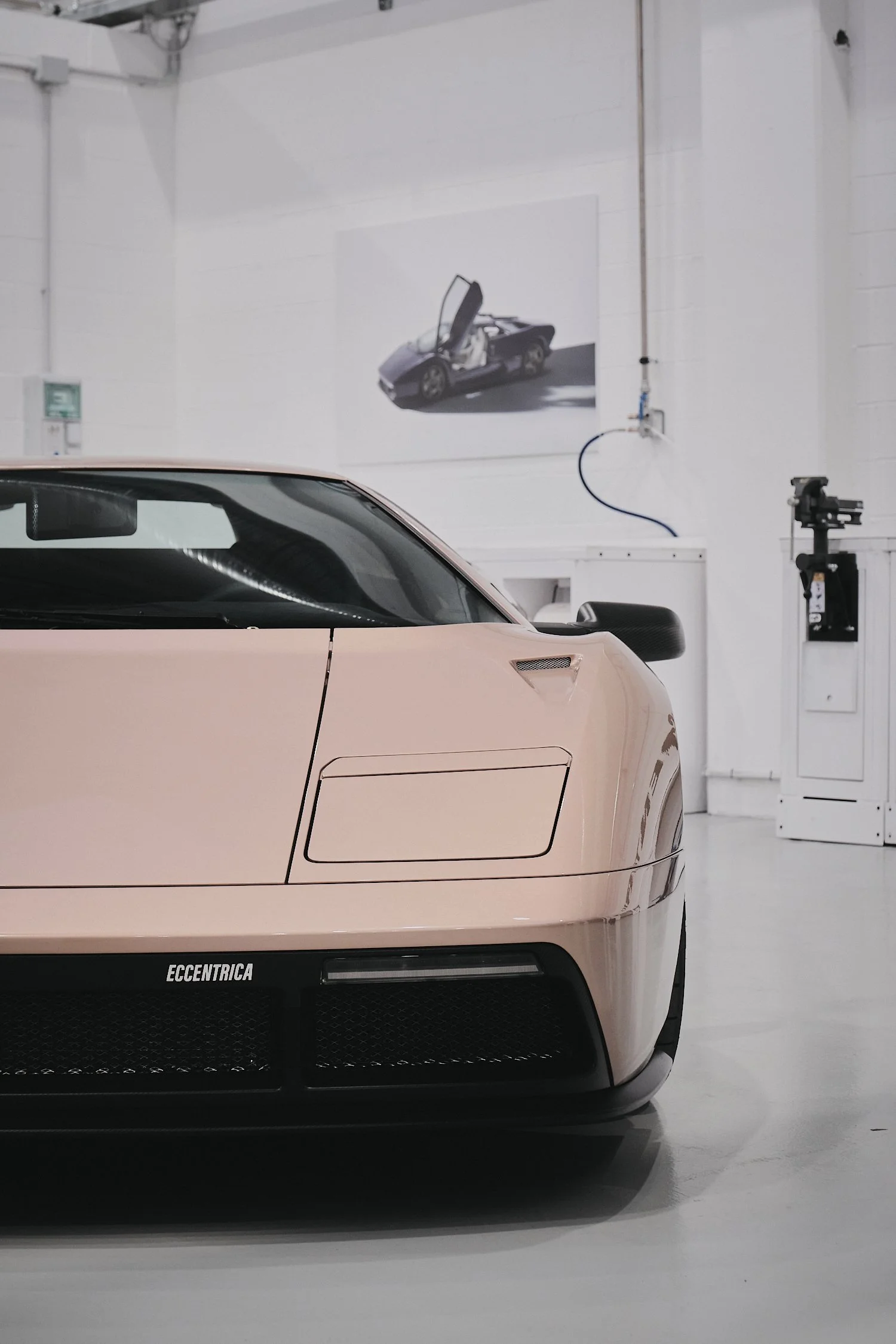 Close-up of the front of a beige sports car labeled "Eccentrica" parked inside a white-walled garage with a framed photo of a car with gullwing doors on the wall behind it.