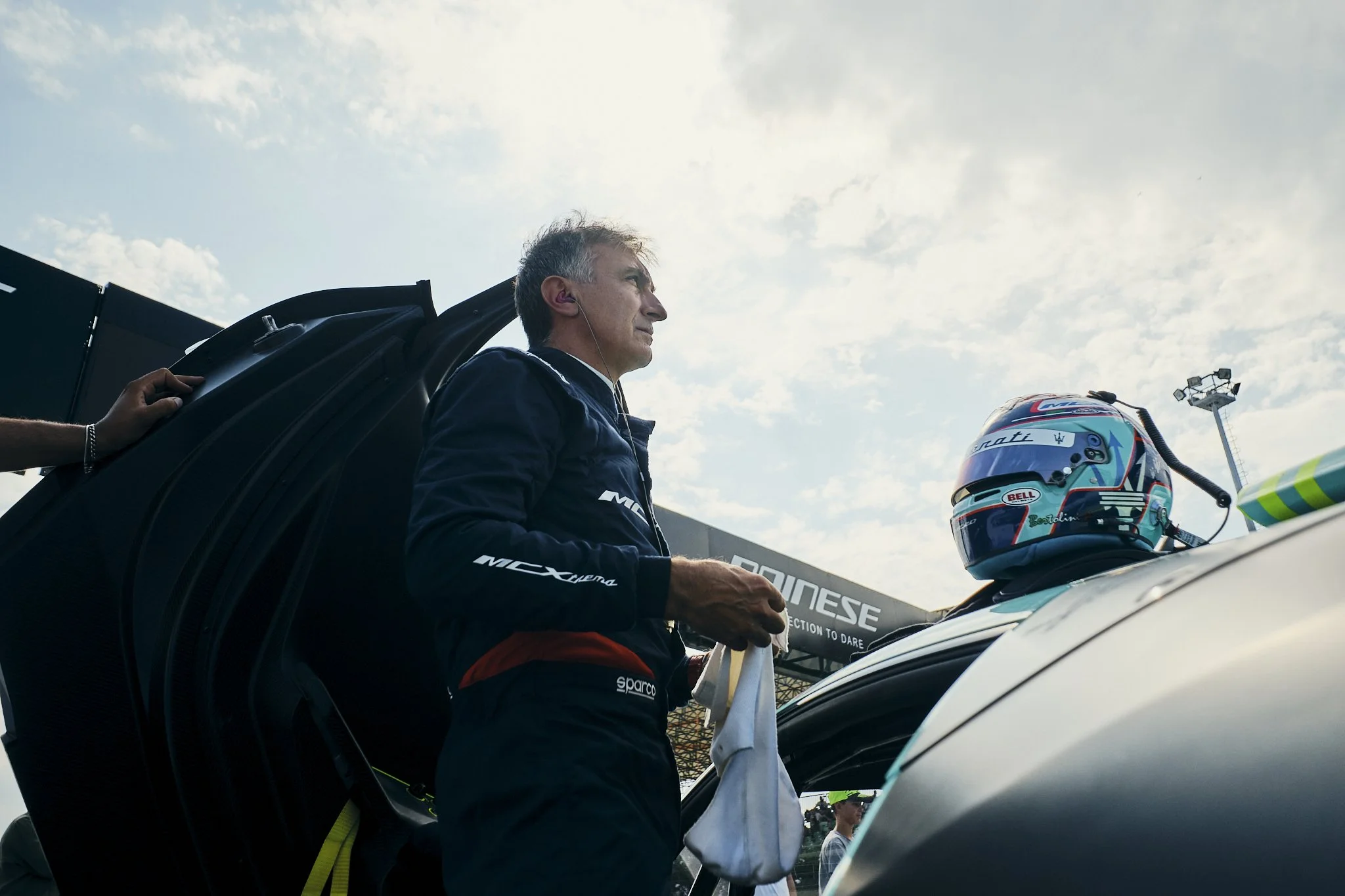 A male race car driver in racing gear standing beside a race car with his helmet placed on the roof, holding a cloth, with a racing track and cloudy sky in the background.