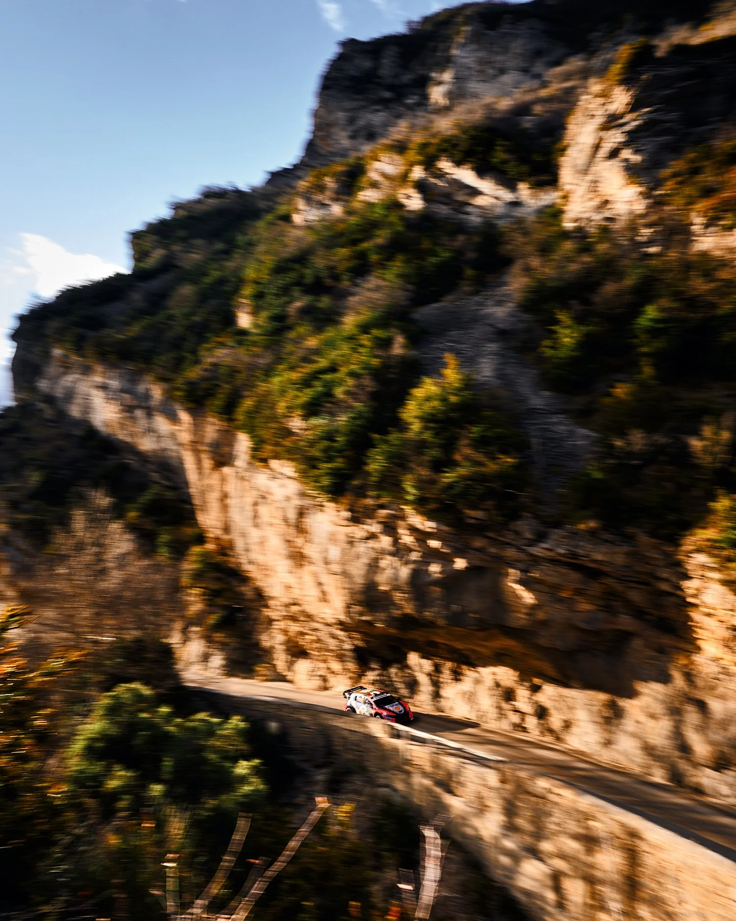 A race car driving on a winding mountain road with large cliffs and green trees in the background.